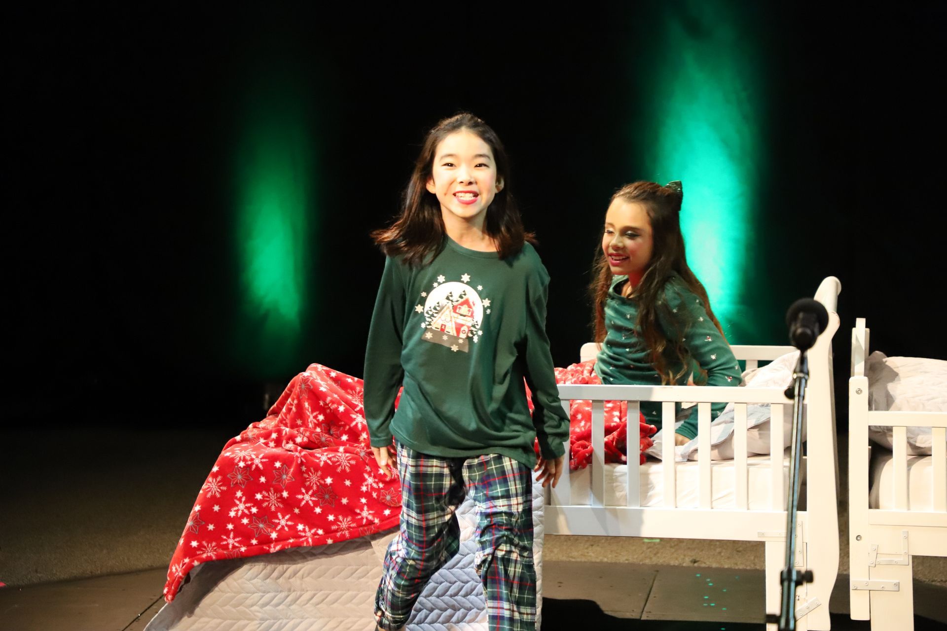 Two young girls are standing next to a white crib on a stage.
