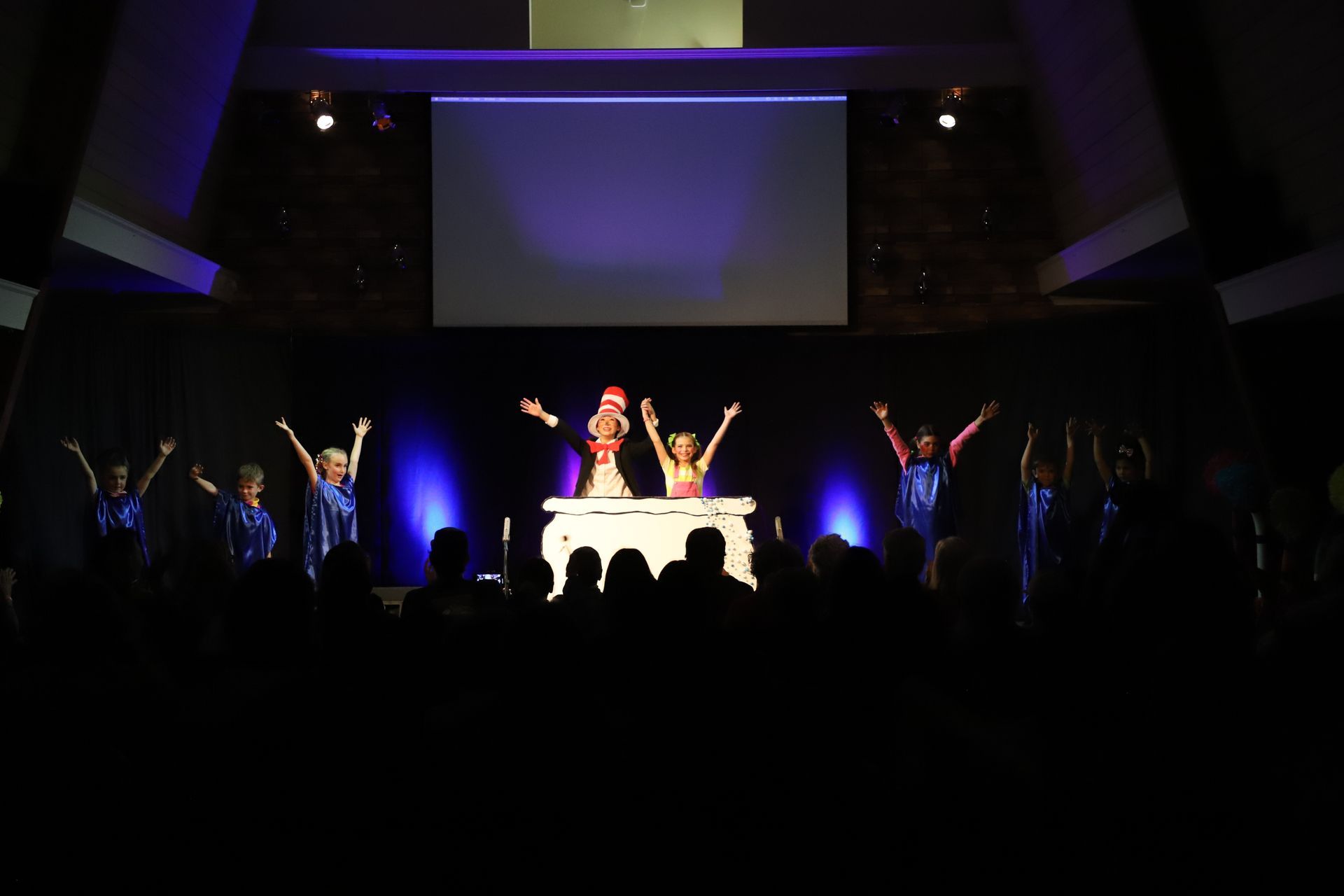 A group of people are dancing on a stage in front of a large screen.