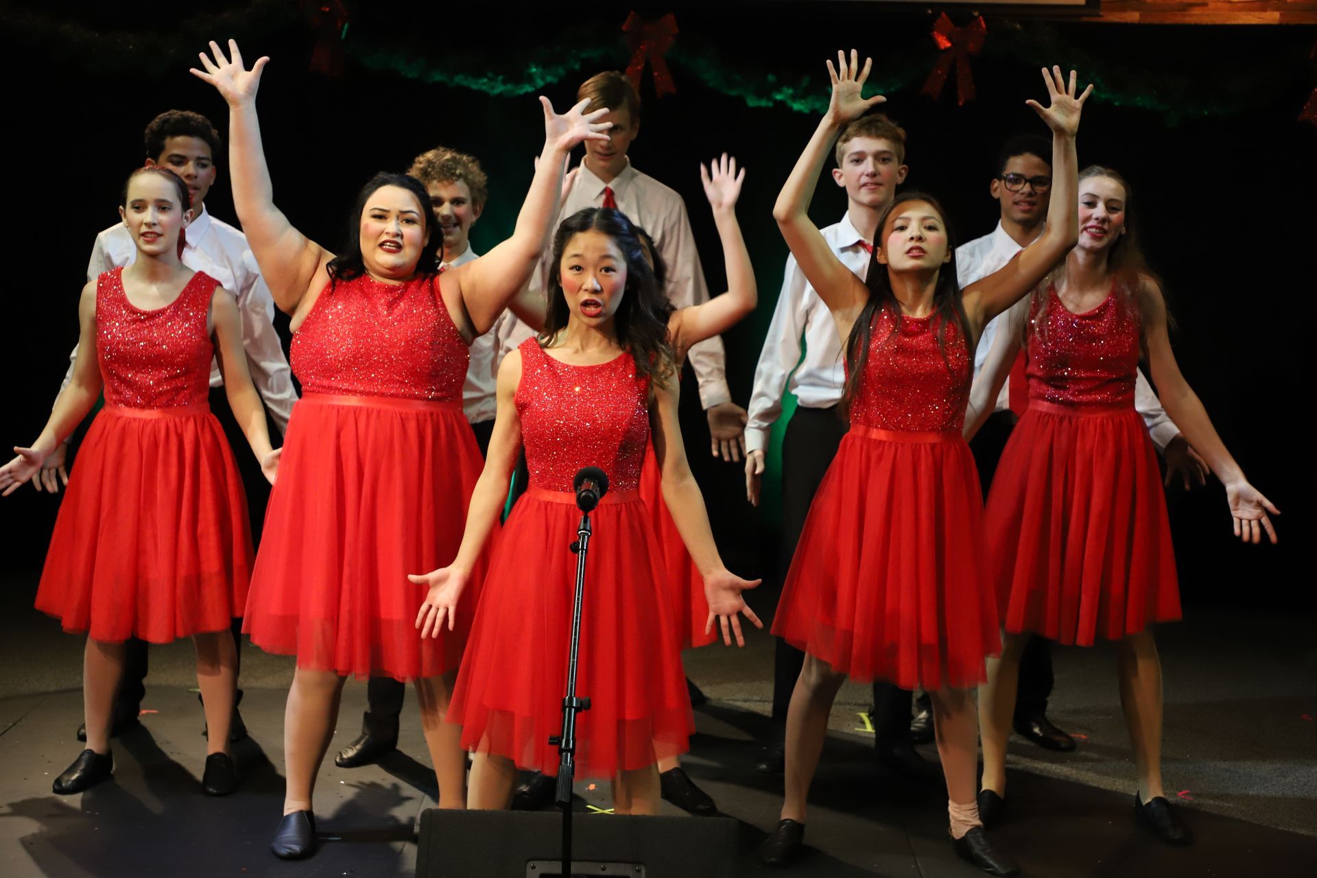 A group of people in red dresses are singing into a microphone on a stage.