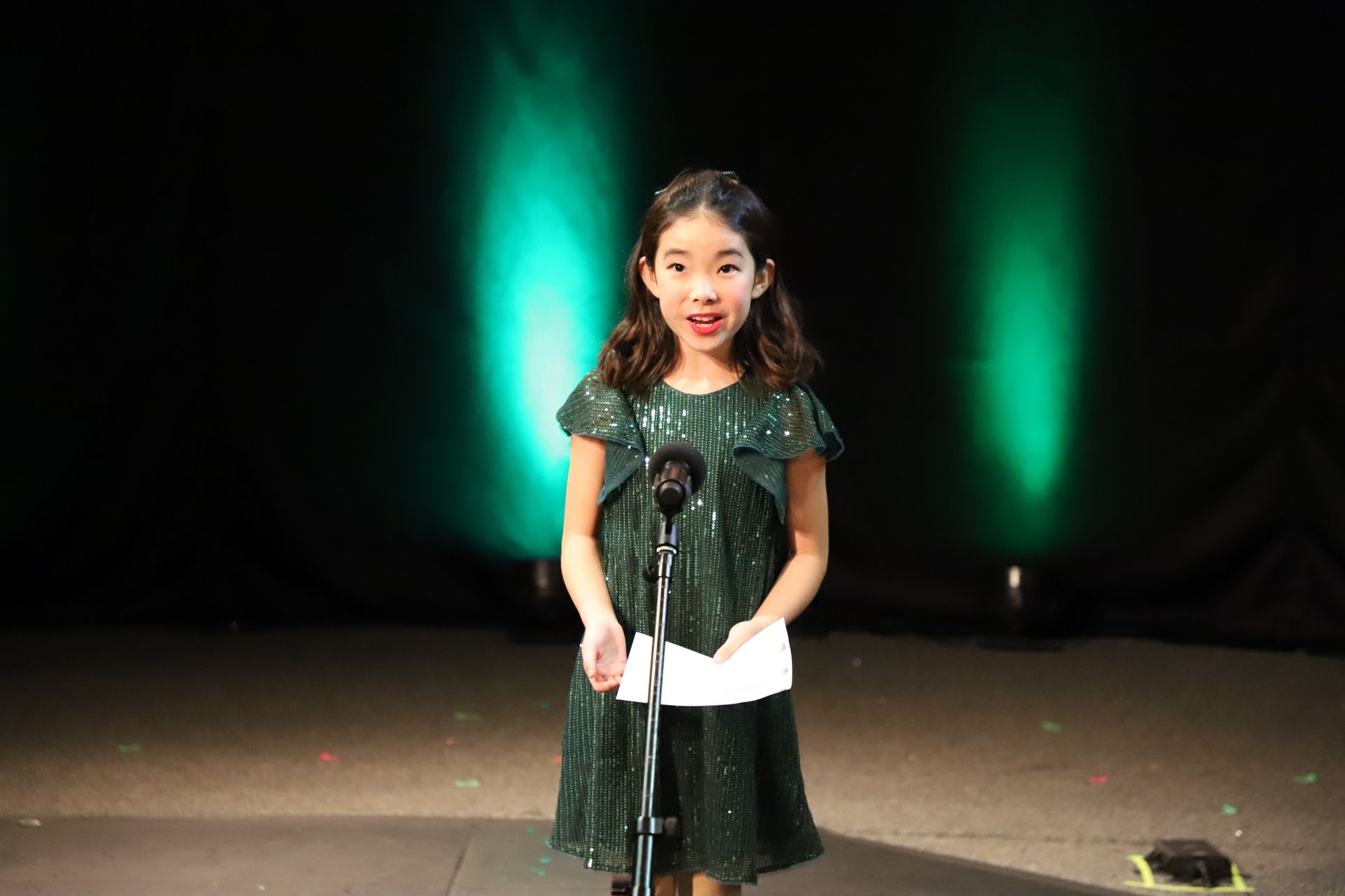 A little girl in a green dress is standing in front of a microphone on a stage.