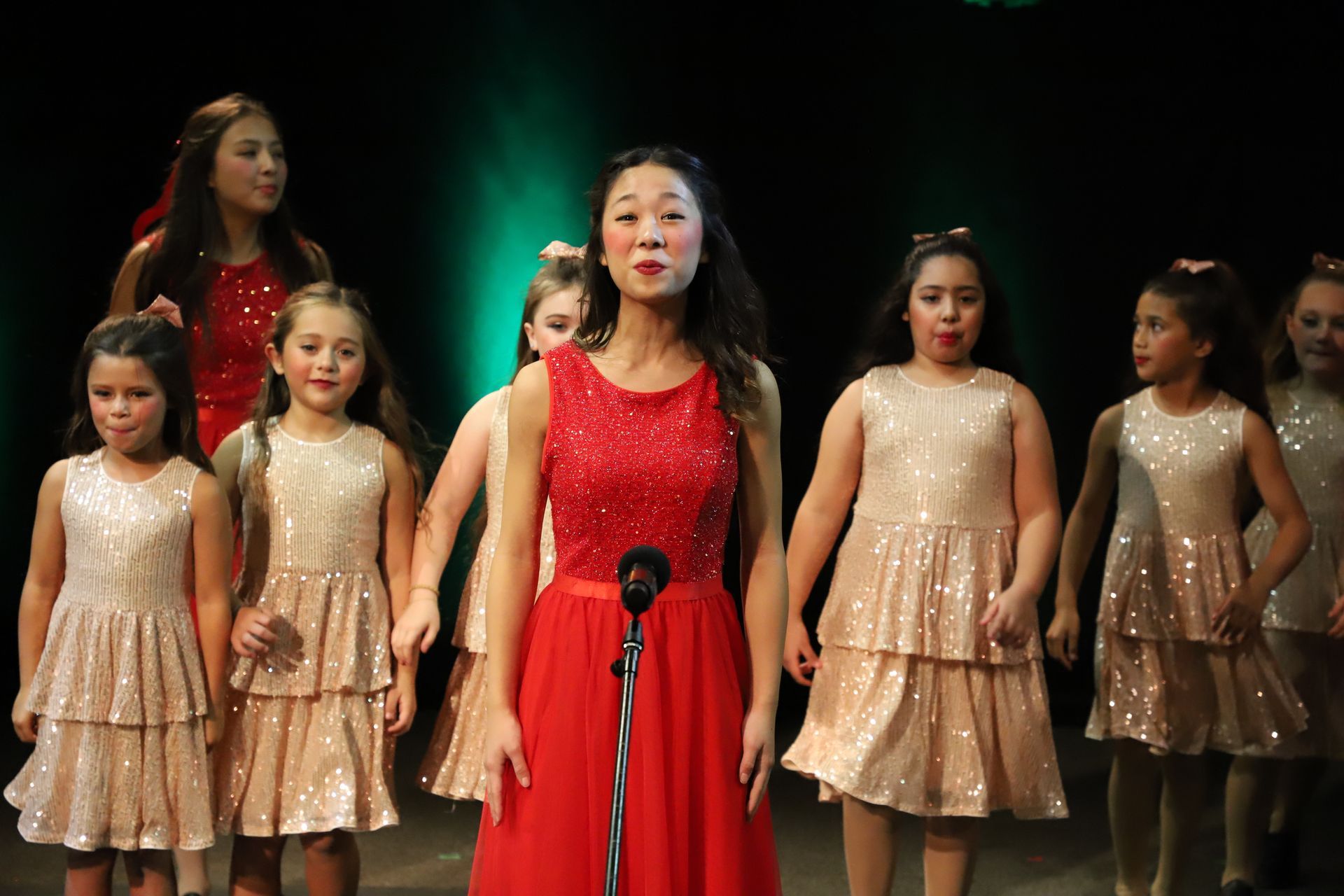 A woman in a red dress is singing into a microphone in front of a group of young girls.