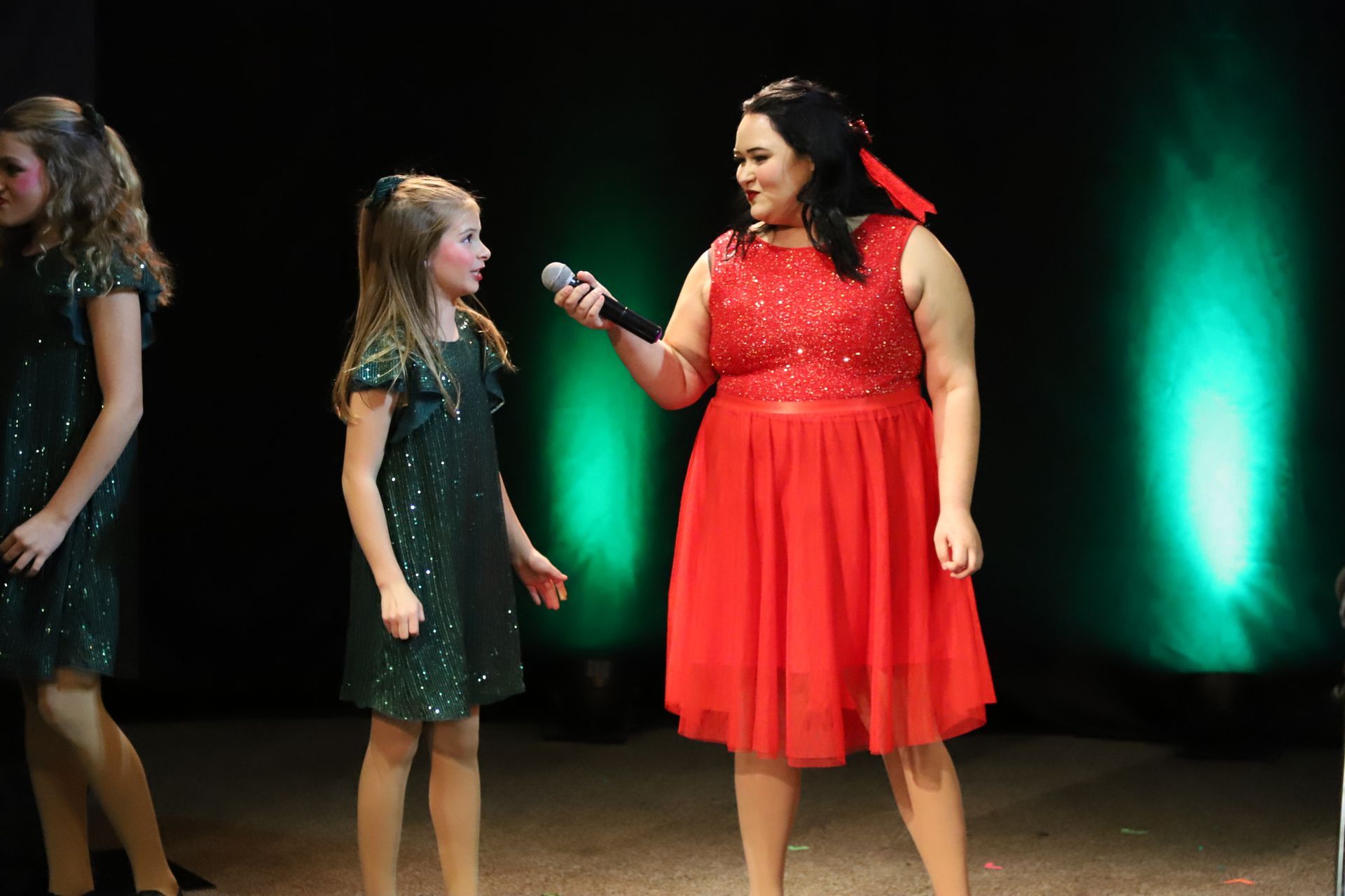 A woman in a red dress is holding a microphone and talking to two young girls.