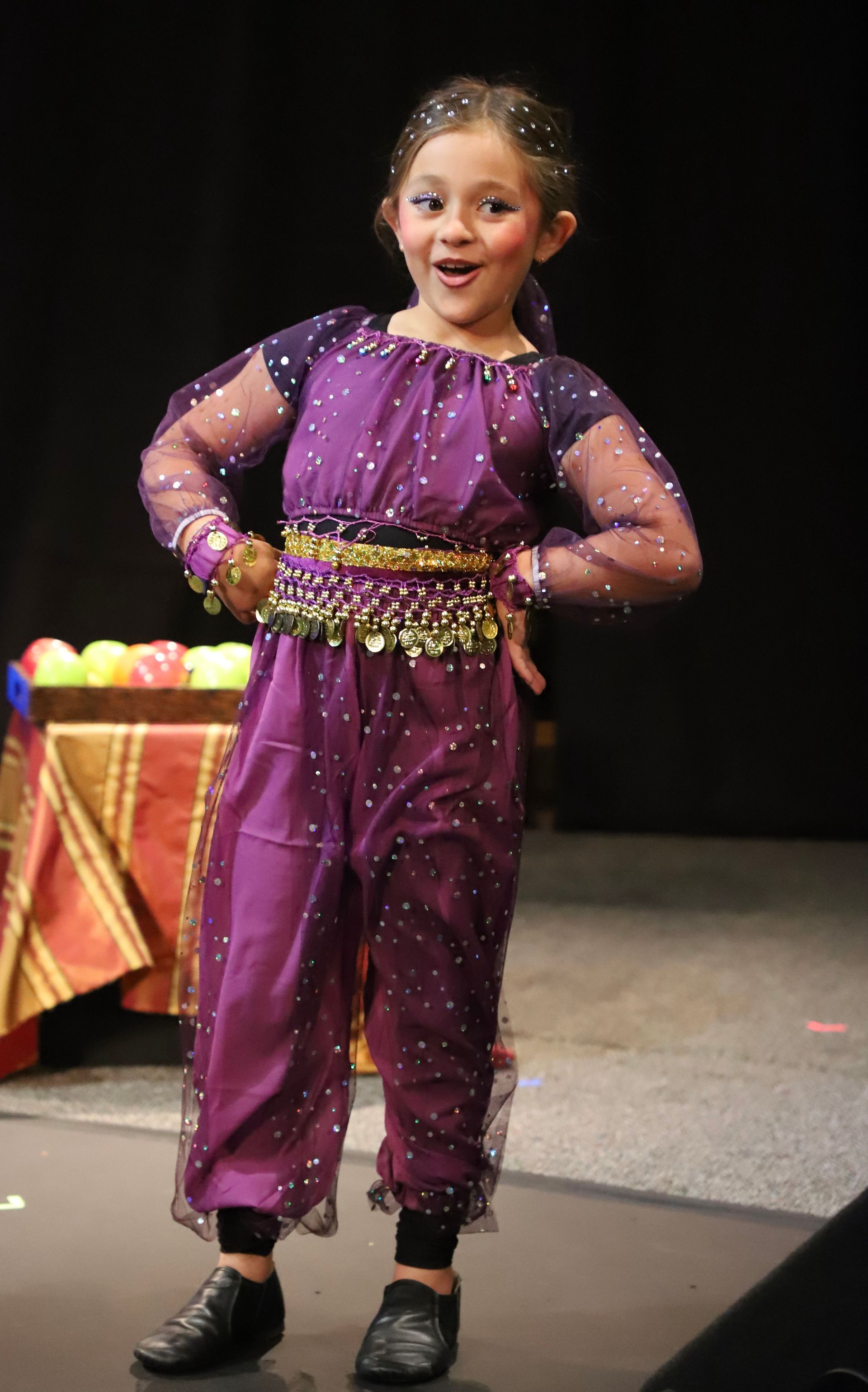 A little girl in a purple dress is standing on a stage.