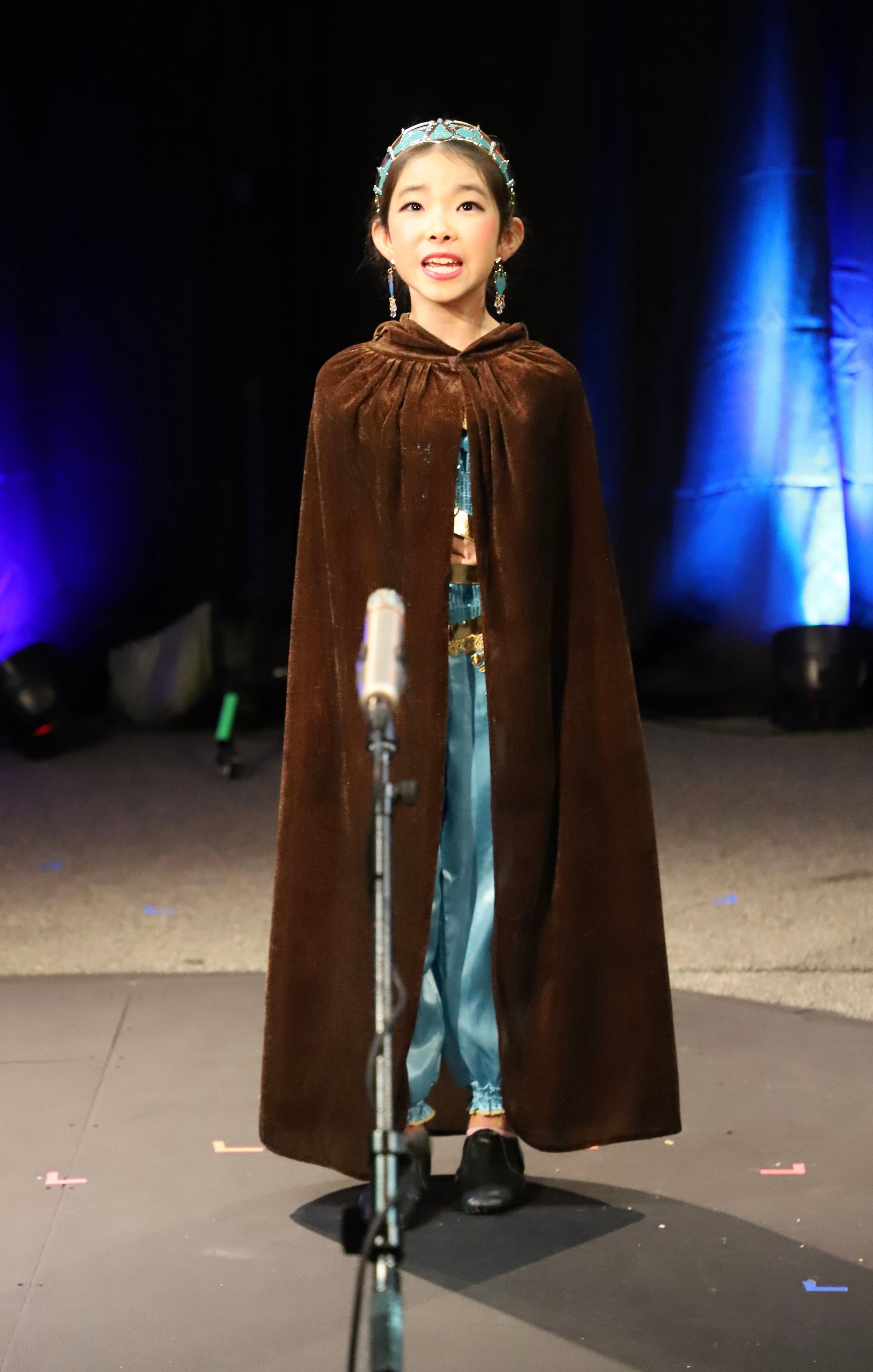 A little girl in a cape is standing in front of a microphone.