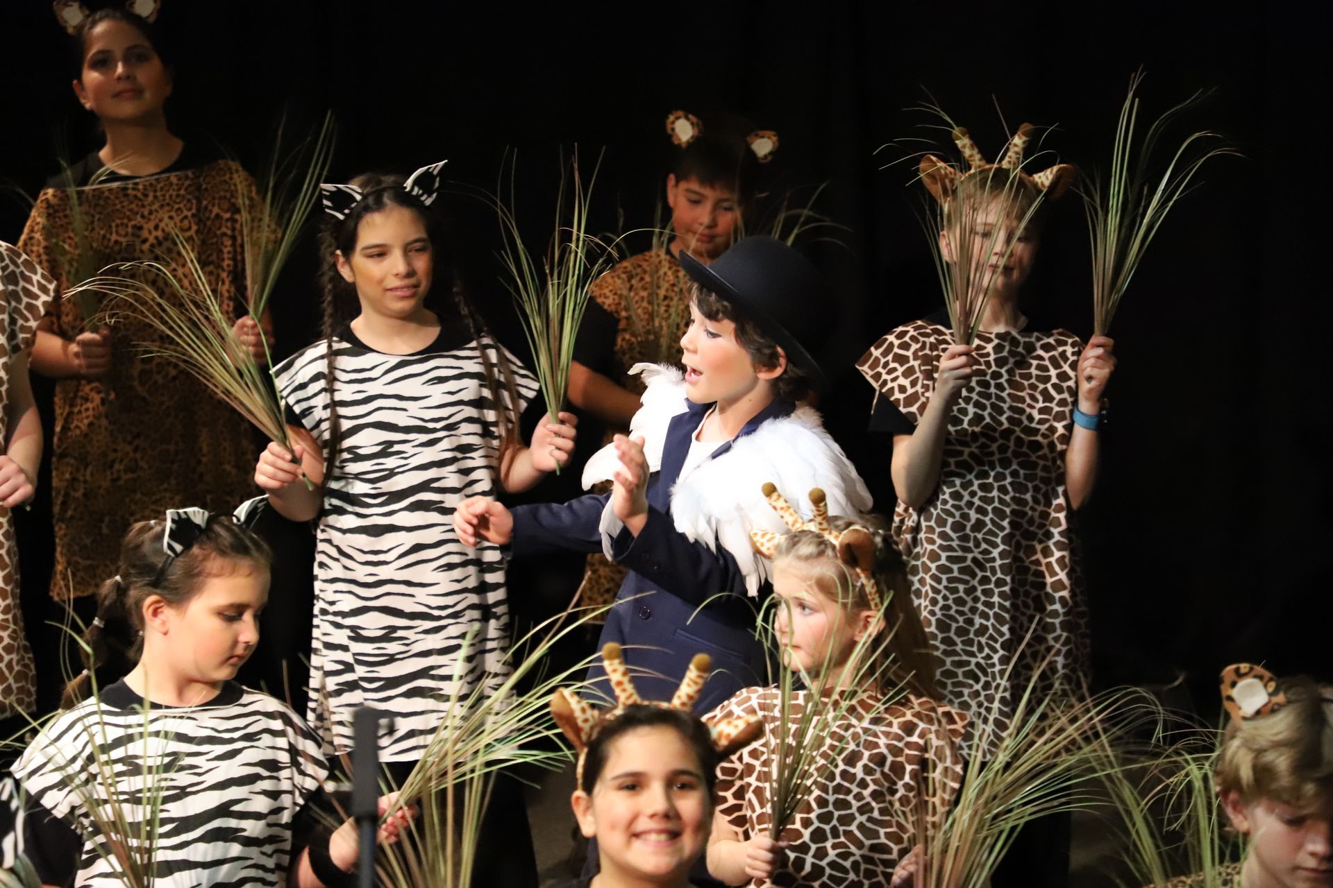 A group of children dressed in zebra and giraffe costumes are holding plants on a stage.