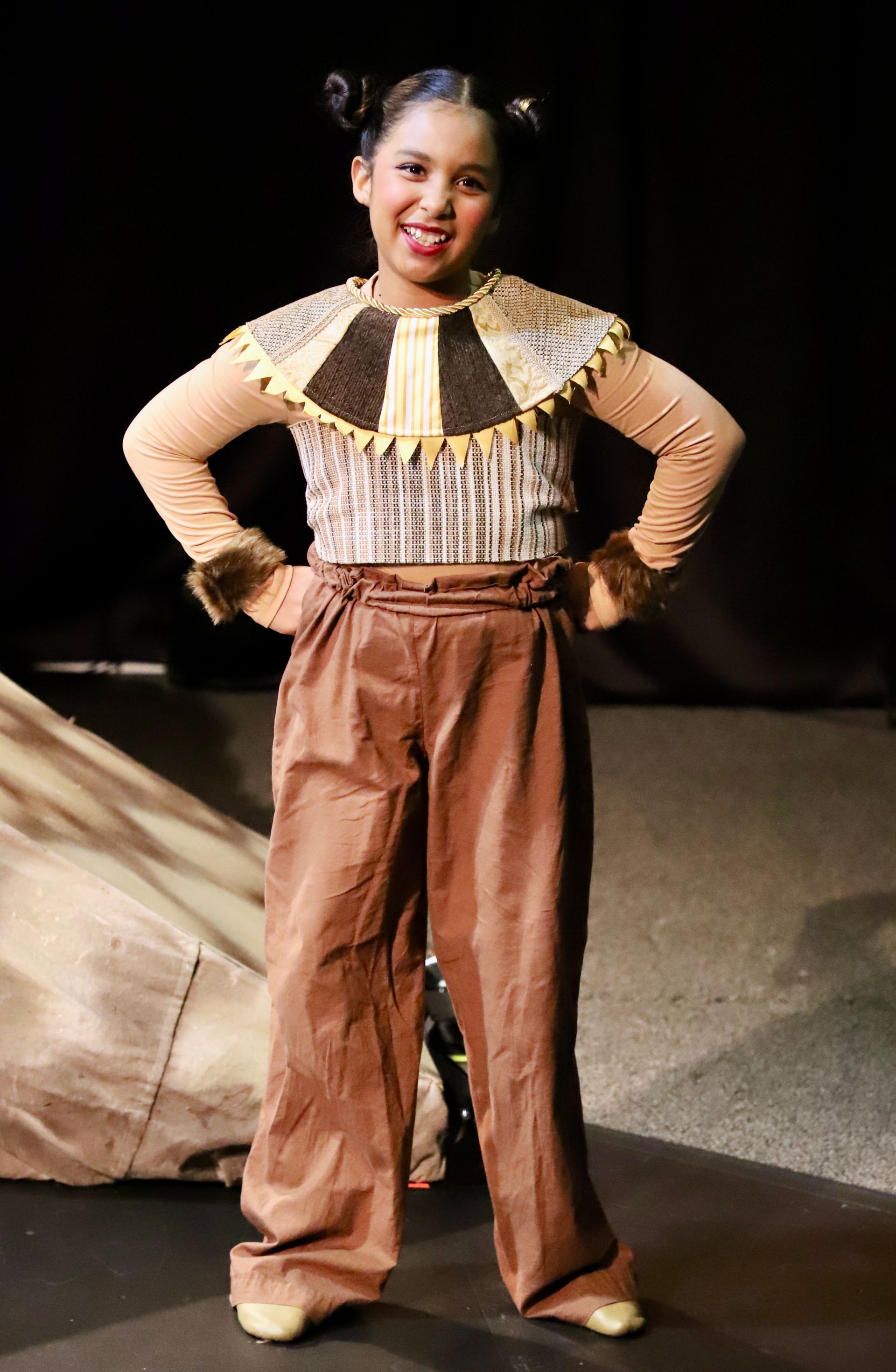 A young girl in a costume is standing on a stage with her hands on her hips.