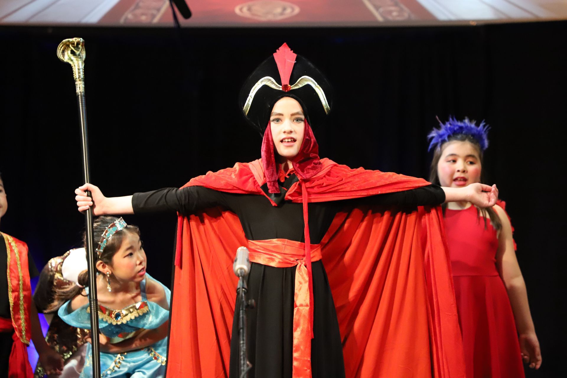 A woman in a red cape is holding a cane in front of a microphone.