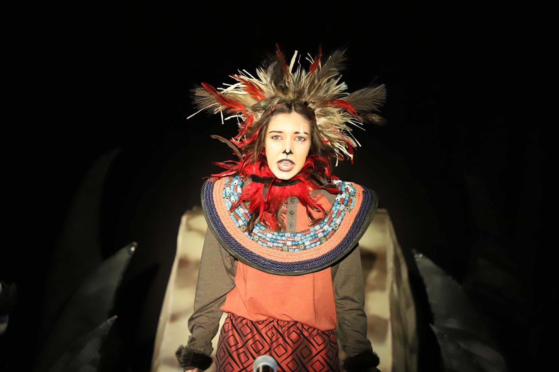 A woman in a costume with feathers on her head is standing in the dark.