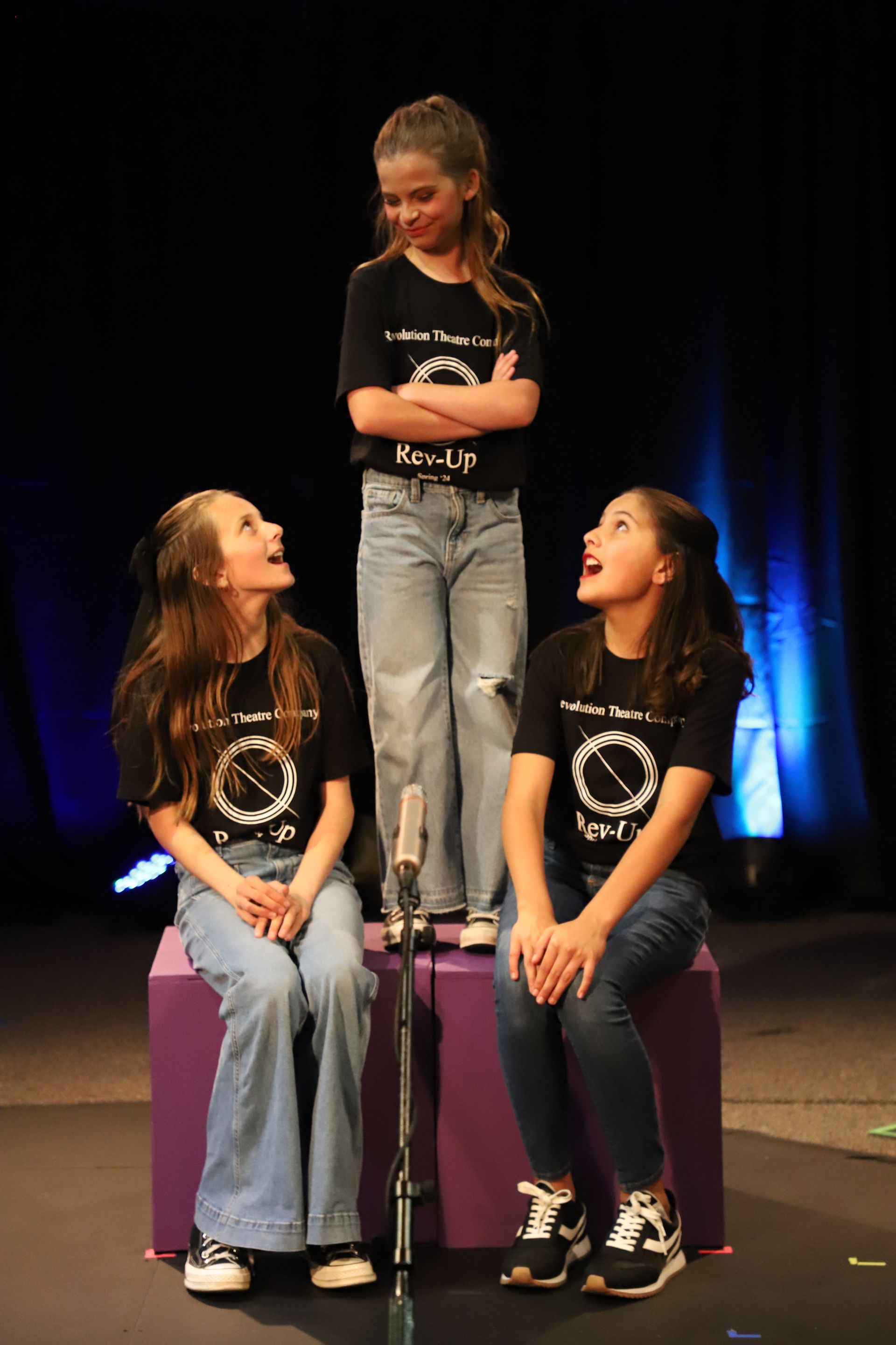 Three young girls are sitting on a purple box on a stage in front of a microphone.