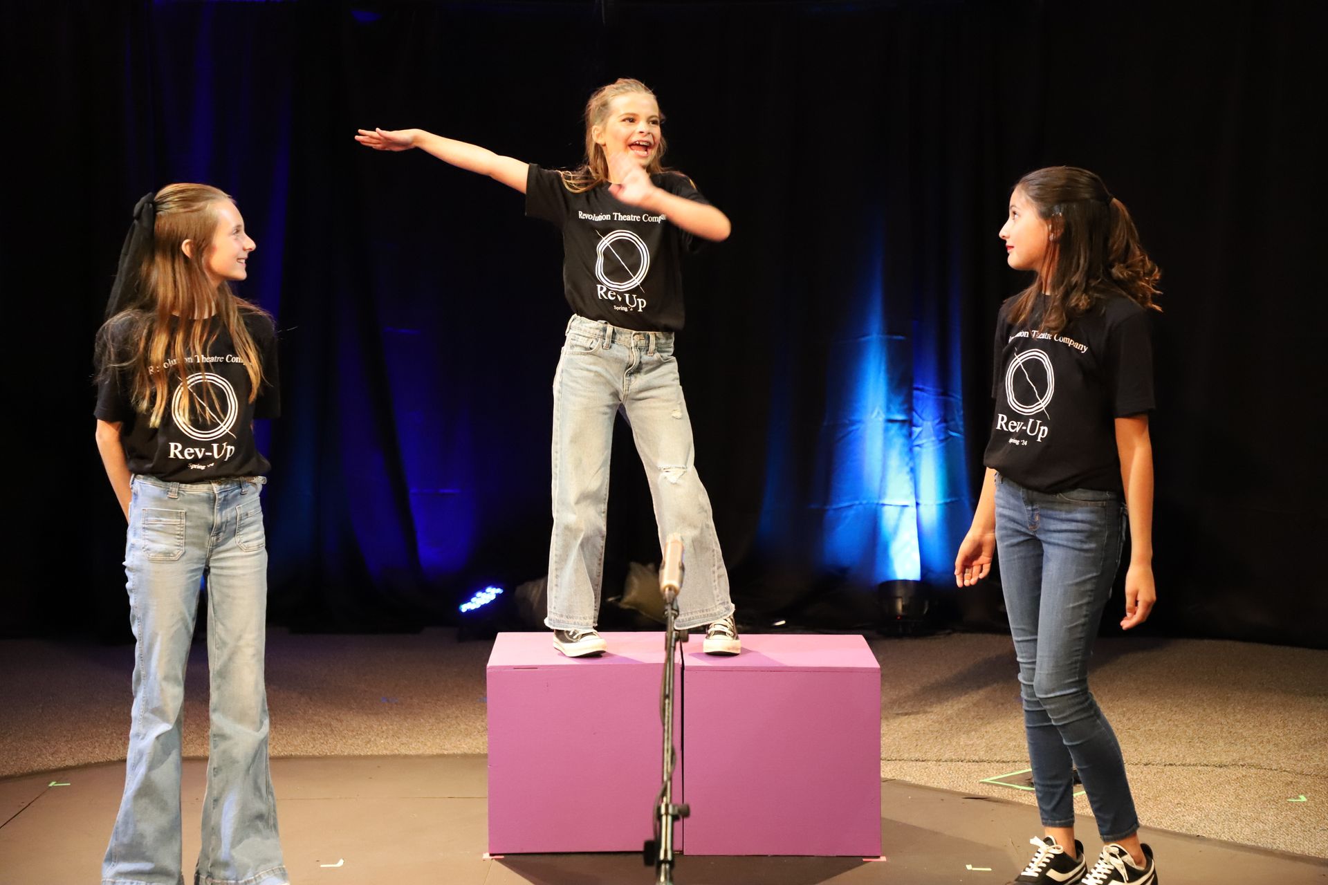 Three young girls are standing on a purple box on a stage.