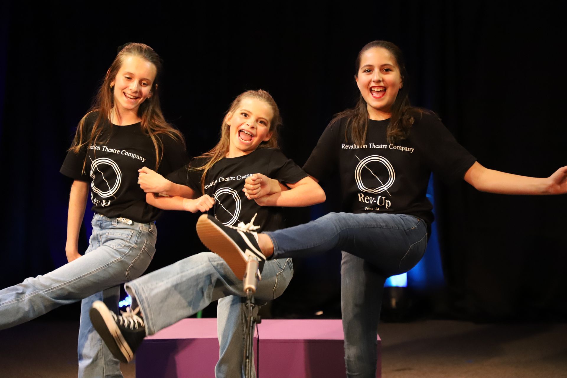 Three young girls wearing black shirts are dancing on a stage.