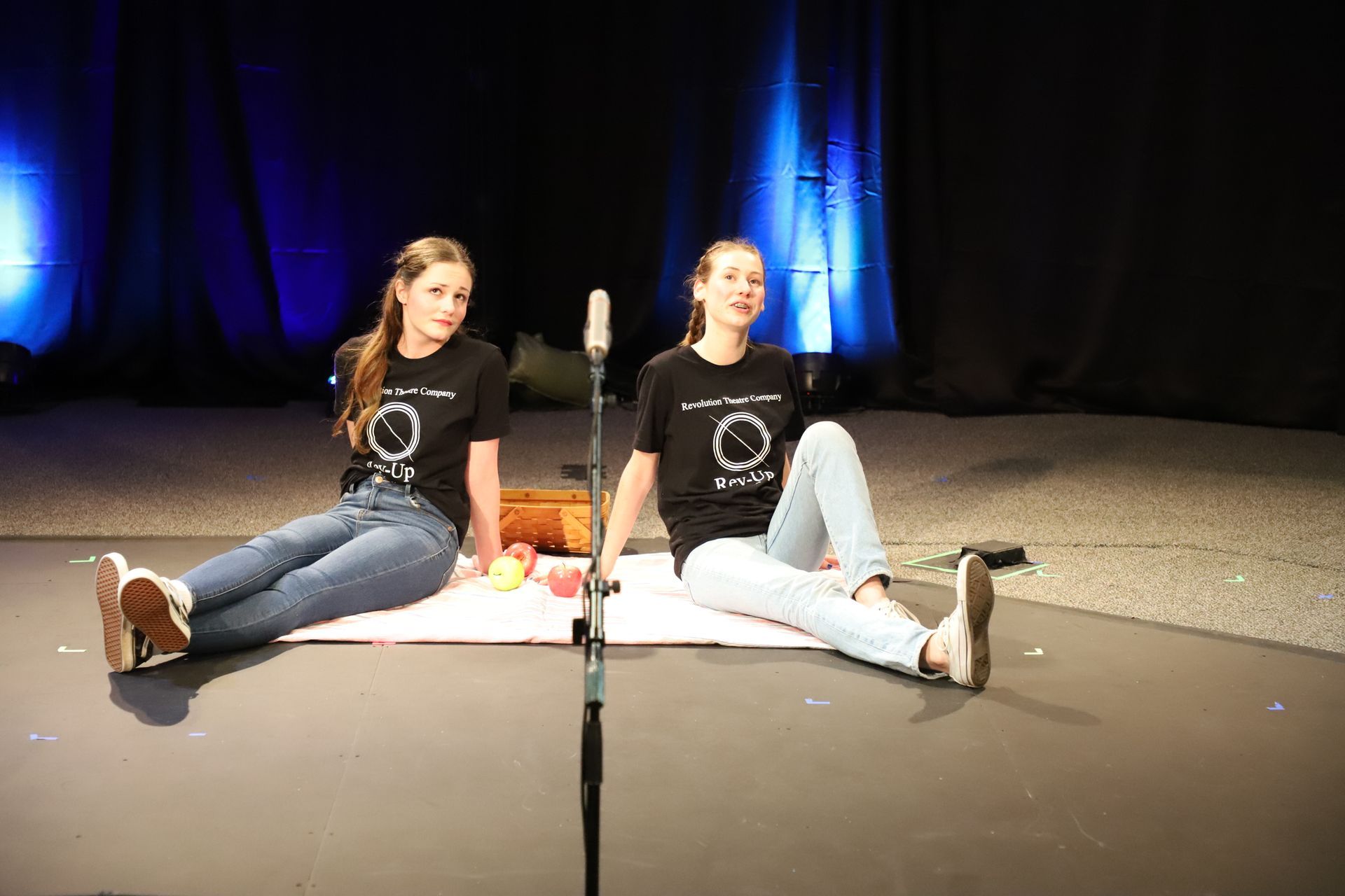 Two girls are sitting next to each other on a stage in front of a microphone.
