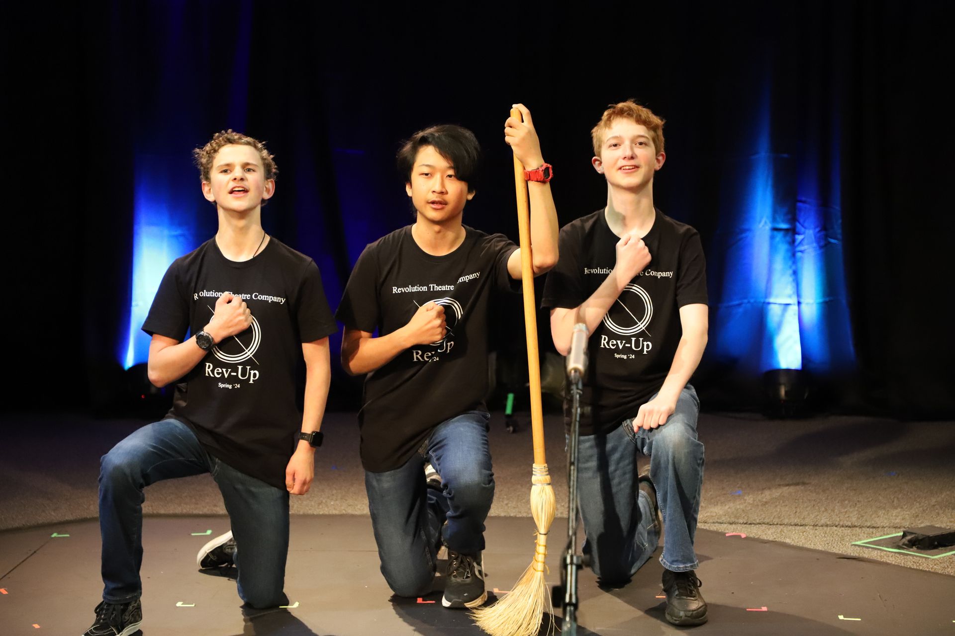 Three young men are kneeling down on a stage holding a broom.