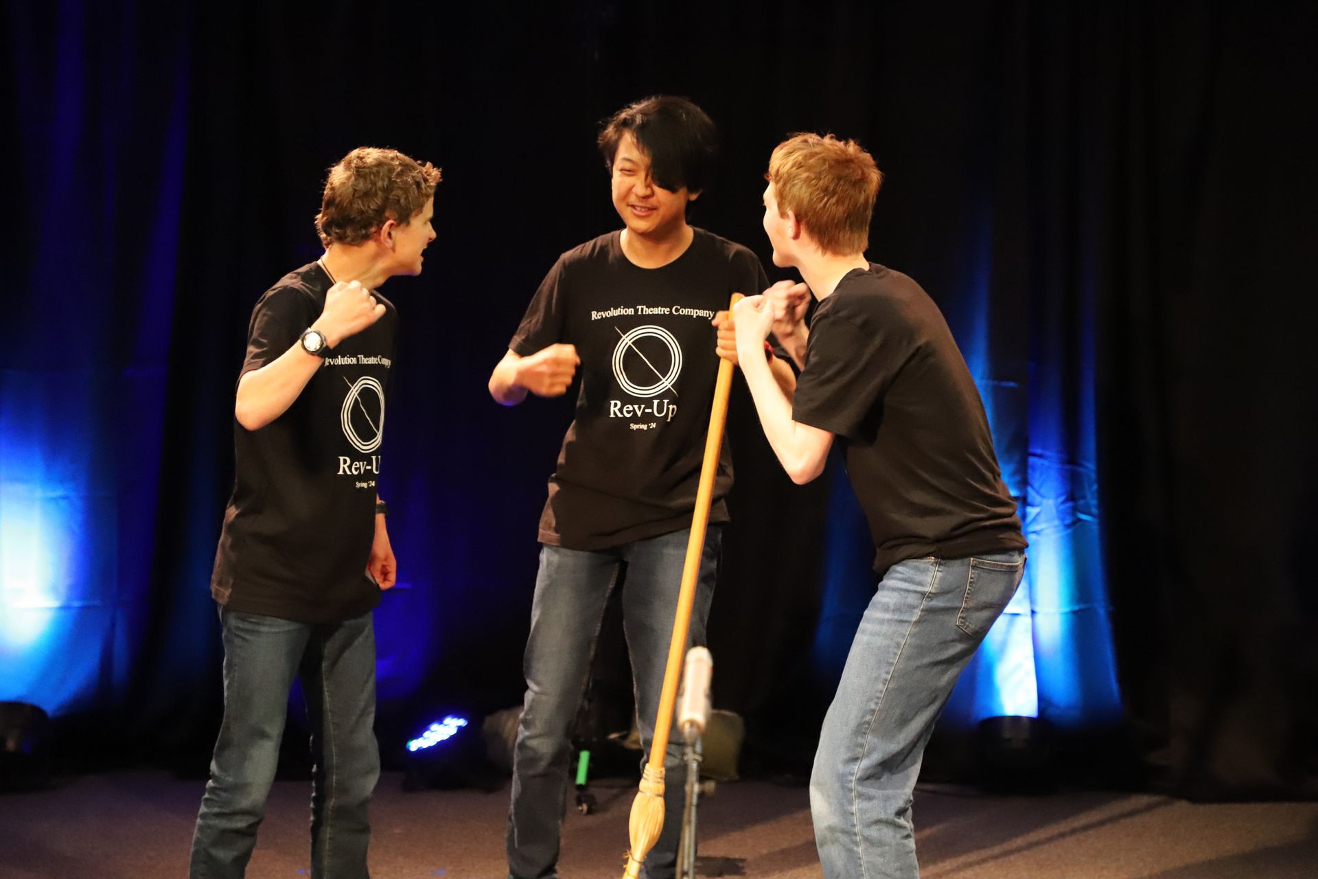 Three young men are standing on a stage and one of them is holding a broom.