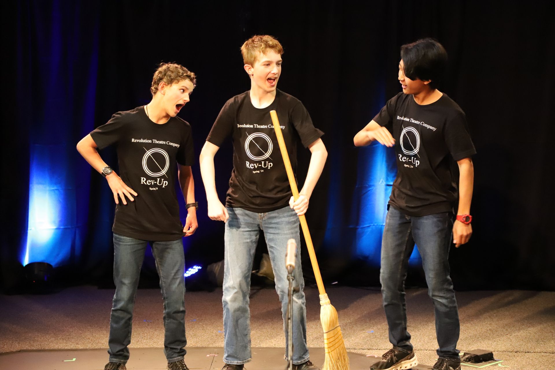 Three young men are standing on a stage holding a broom.