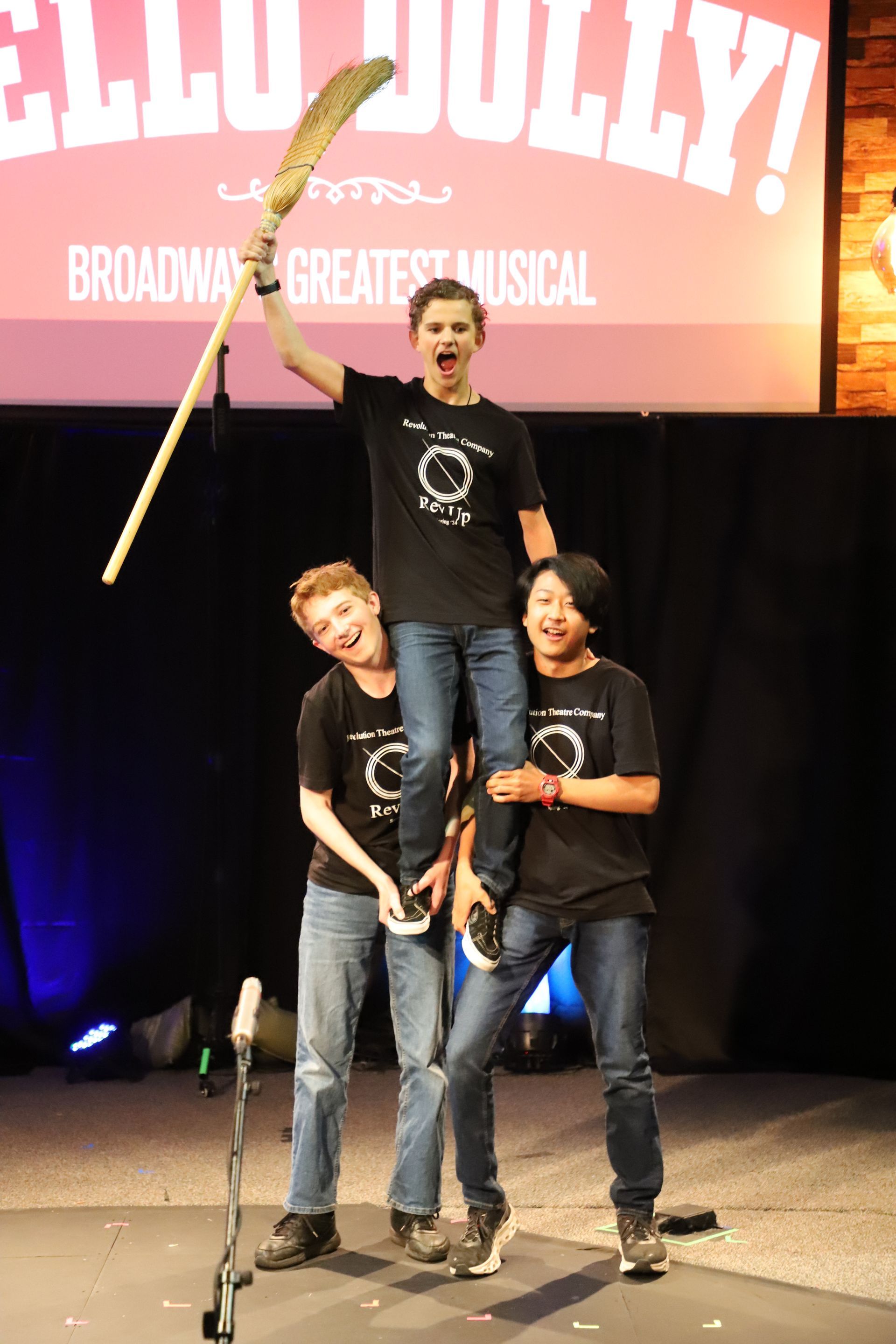 Three young men are standing on a stage holding a broom.