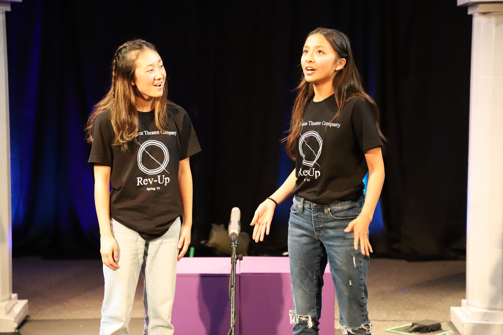 Two girls are standing next to each other on a stage in front of a microphone.