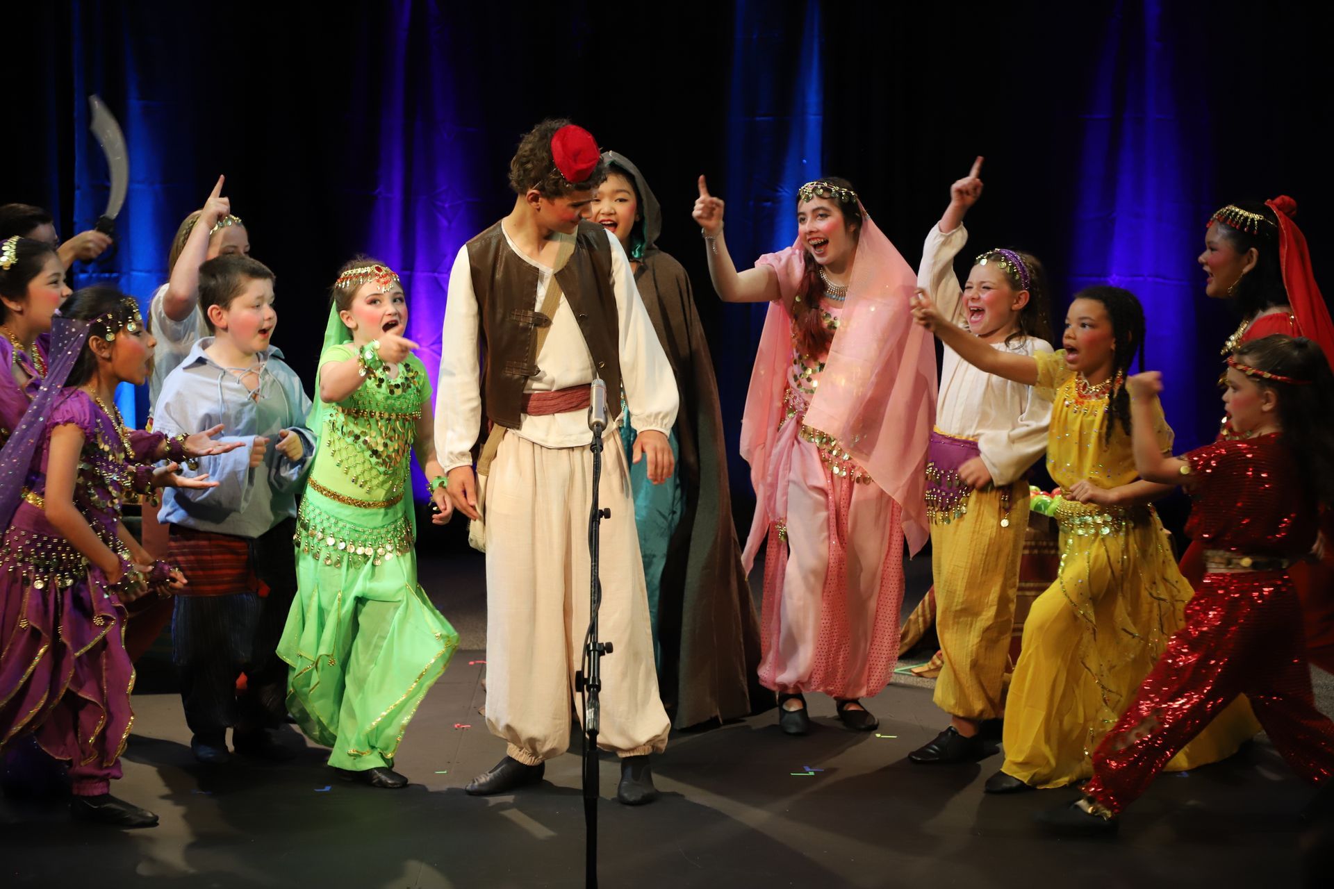A group of children are dancing on a stage while a man sings into a microphone.