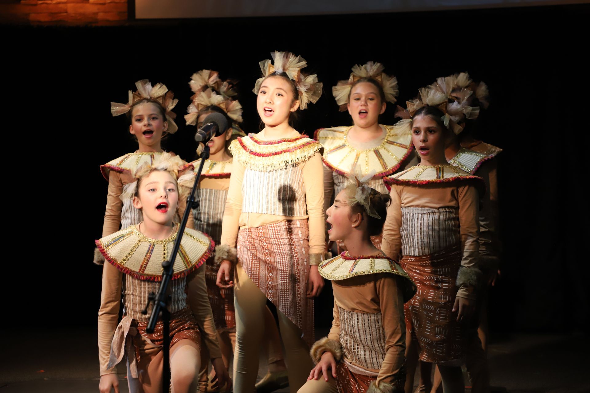 A group of young girls in costume are singing into microphones on a stage.