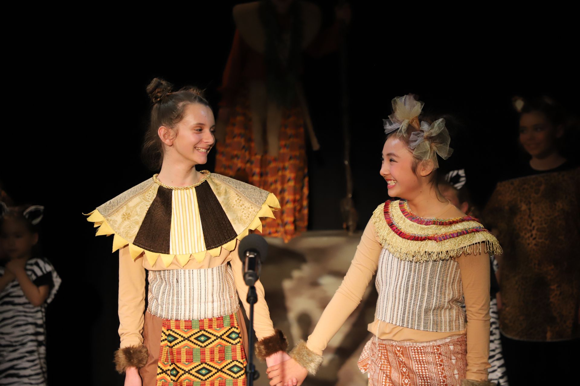 Two young girls in costume are holding hands on a stage.