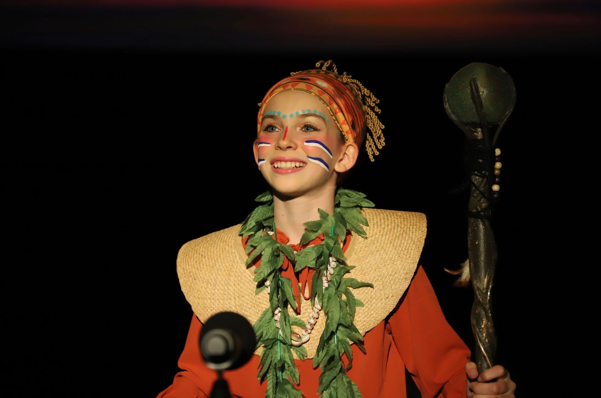 A young boy in a hawaiian costume is holding a stick and smiling.