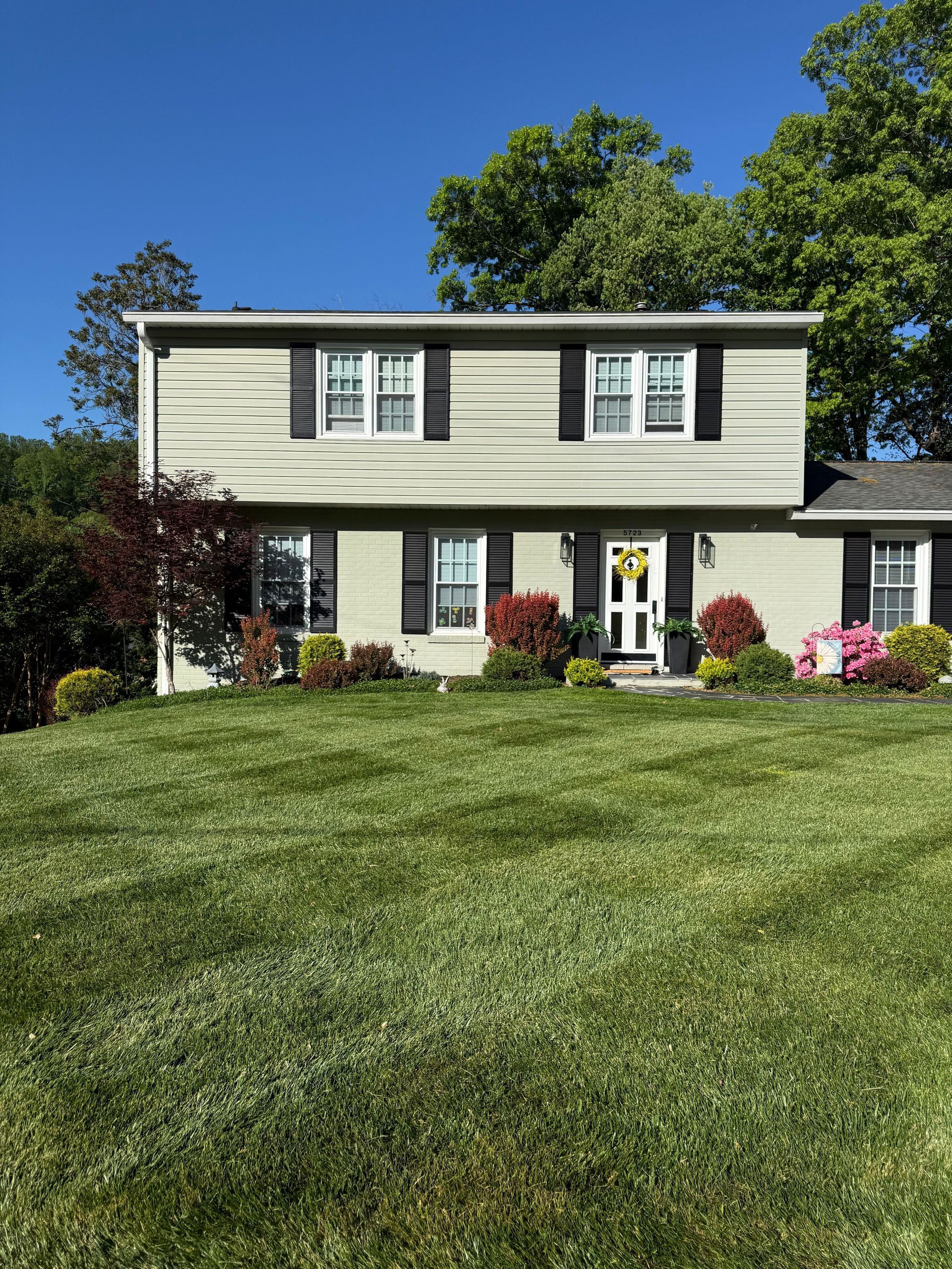 Two-story beige house with white trim, black shutters, and a large green lawn under a clear blue sky