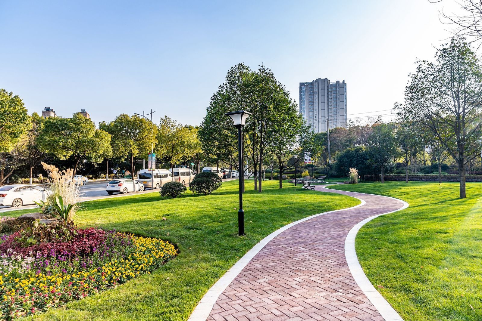 Curved brick path through a green park with flowers, trees, and a tall building in the background.