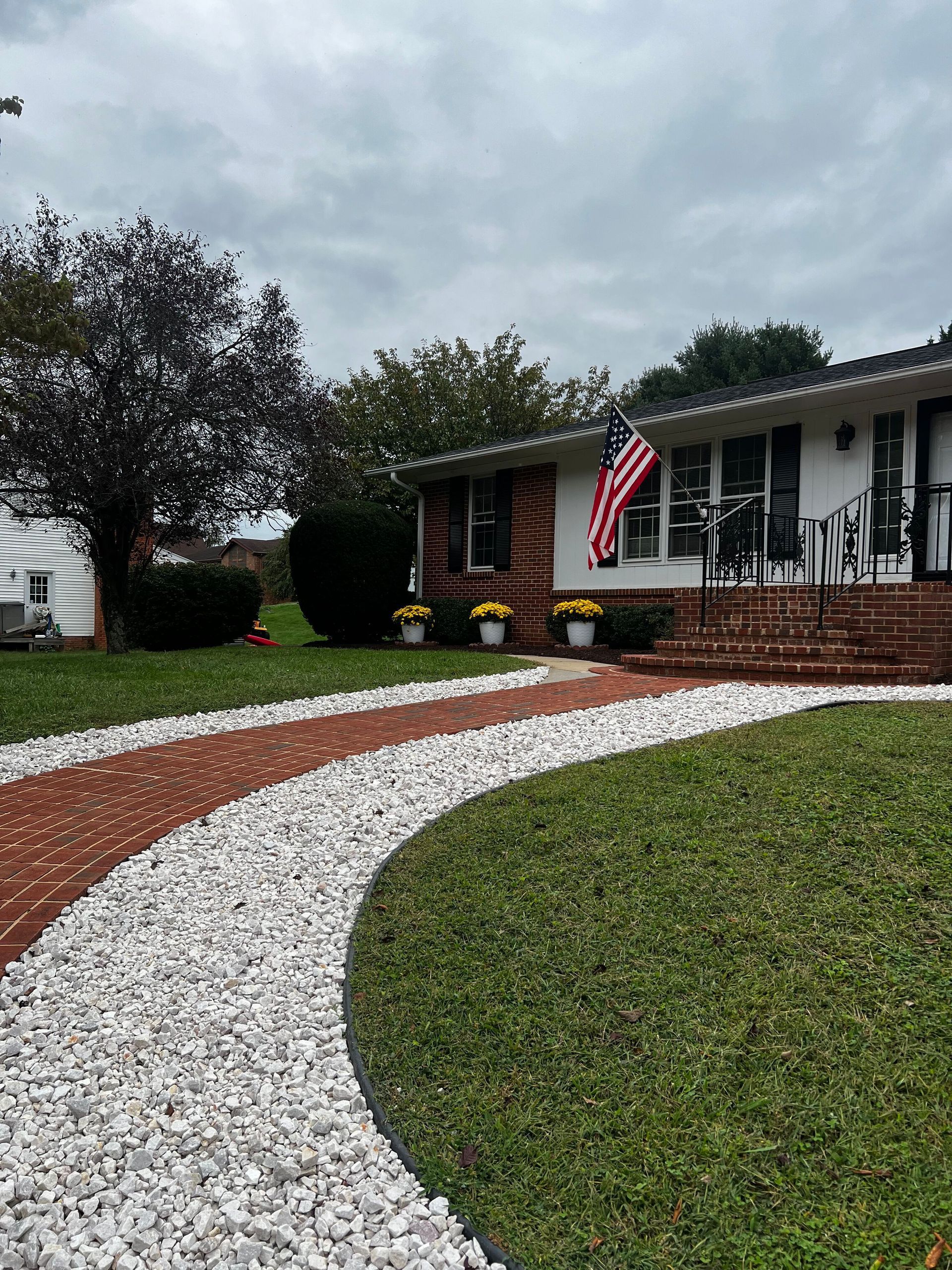 Front yard with white gravel path, green lawn, brick porch, and a house with an American flag under cloudy skies