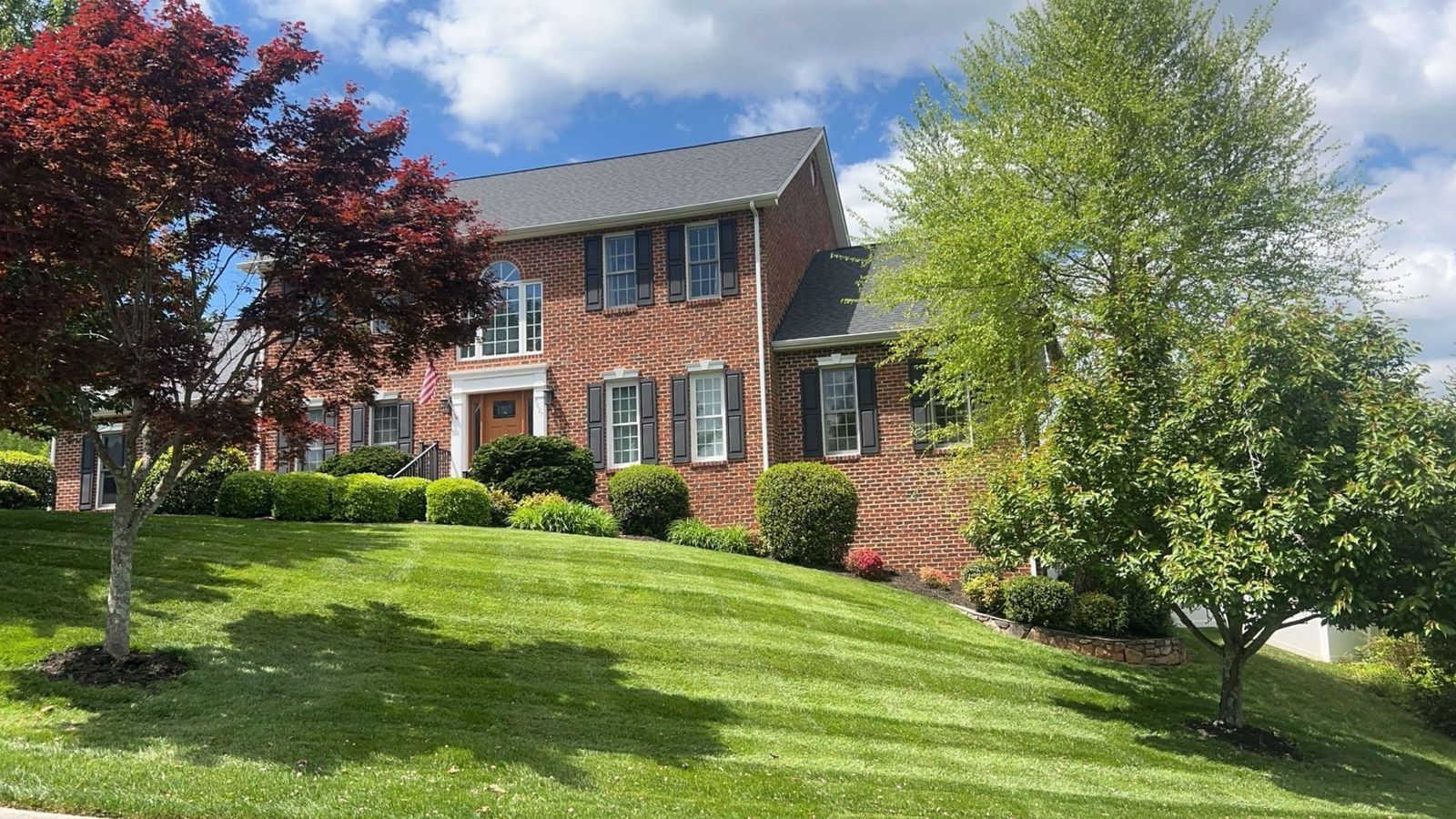 Brick two-story house on a green lawn with trees under a blue sky.