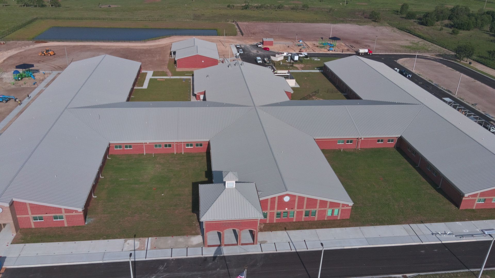Aerial view of a red brick school building with a metal roof. A playground and pond are nearby.