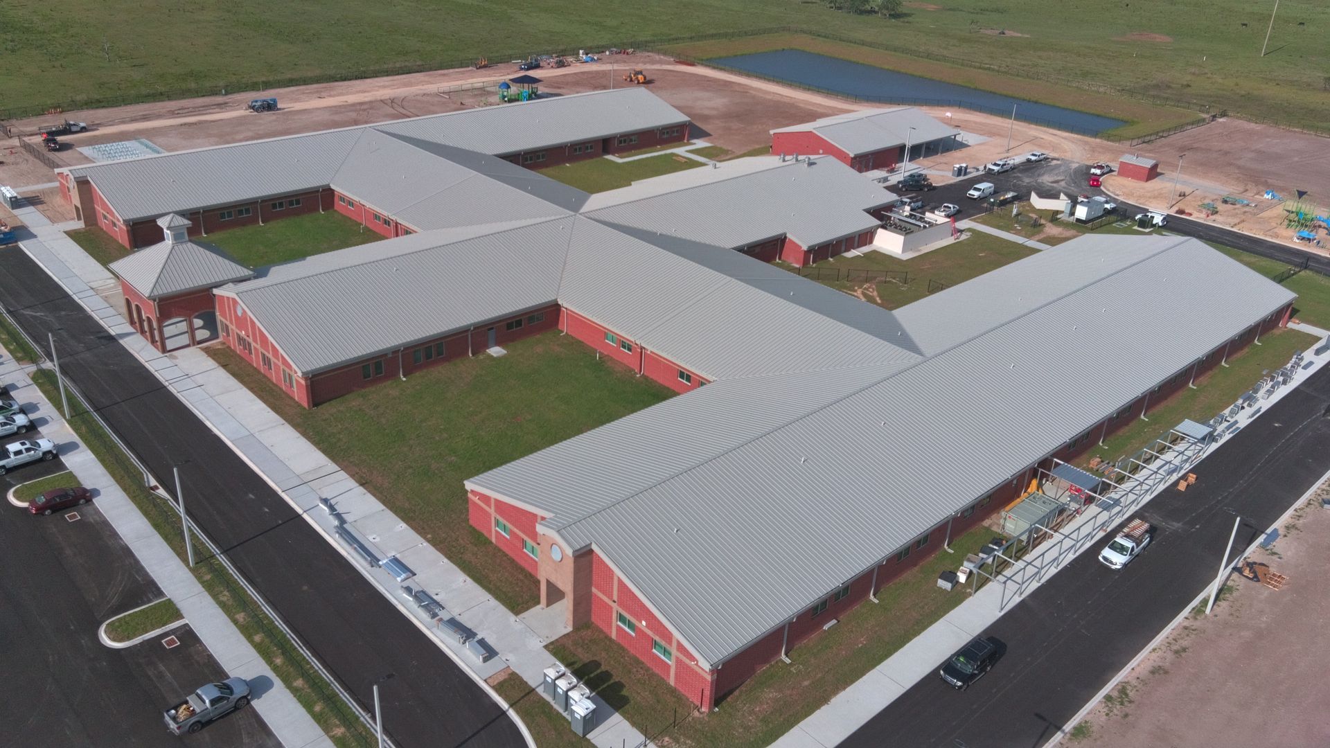 Aerial view of red buildings with silver roofs arranged in a multi-winged complex, surrounded by green grass and a parking lot.