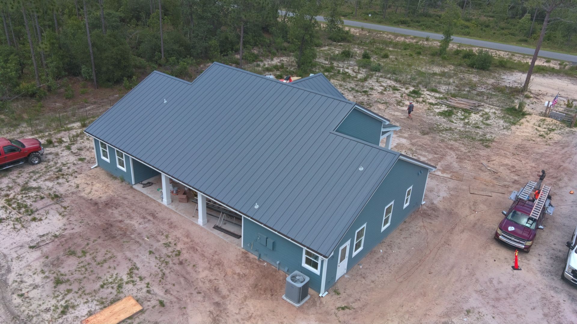 Aerial view of a teal-colored house under construction with a metal roof, surrounded by dirt and vehicles.