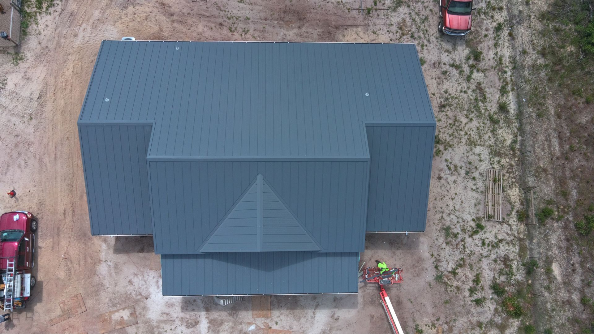 Overhead view of a house with a dark gray metal roof, construction vehicles, and a worker on a lift.