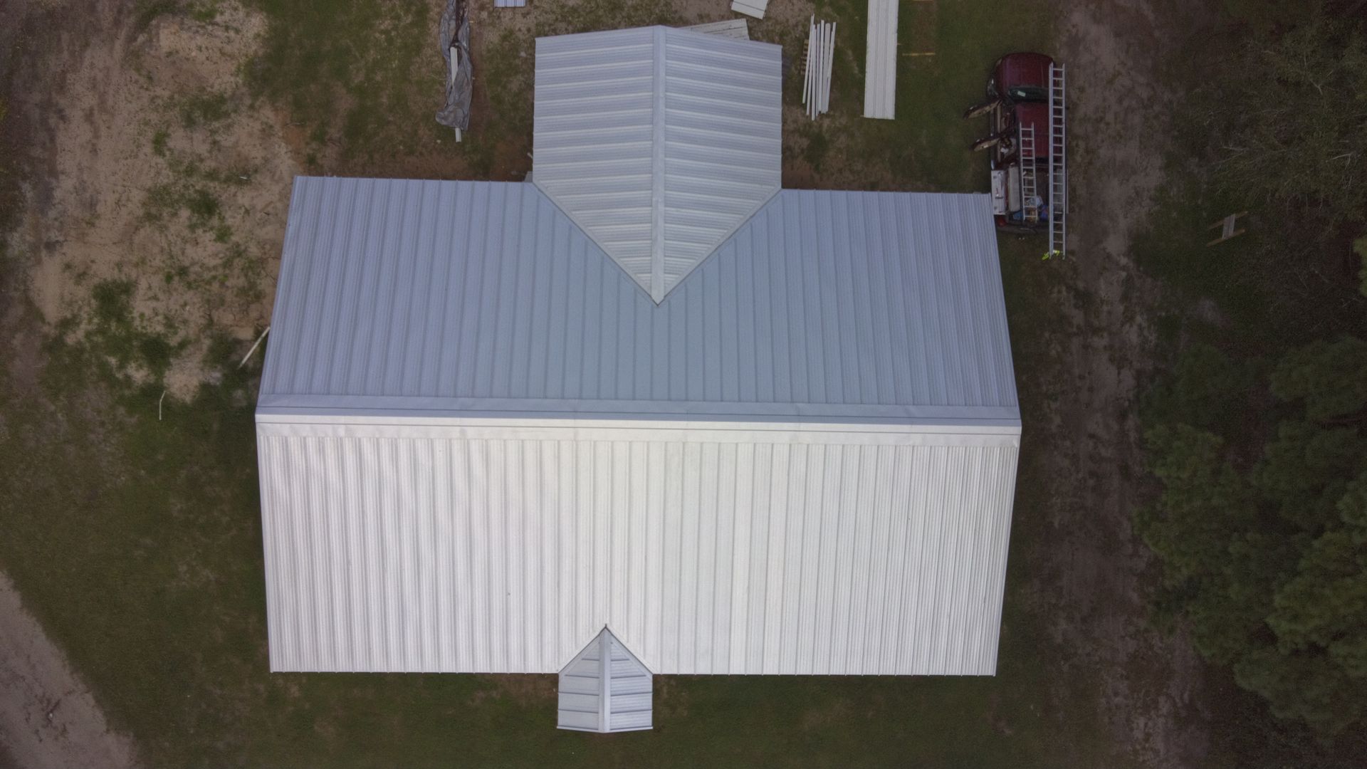 Overhead view of a metal-roofed building with a centered extension. A ladder and other materials sit nearby.