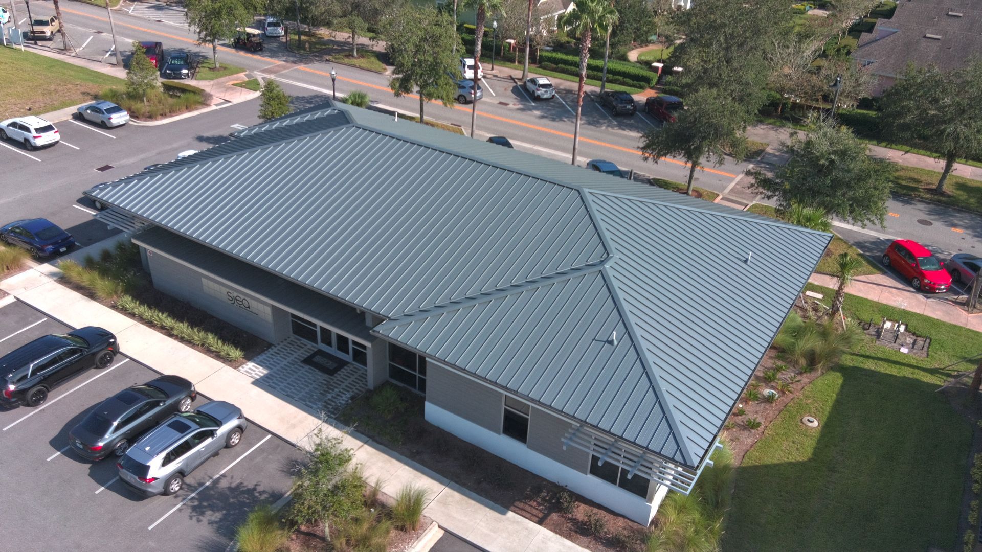 An aerial view of a building with a gray metal roof, surrounded by parking and a street.