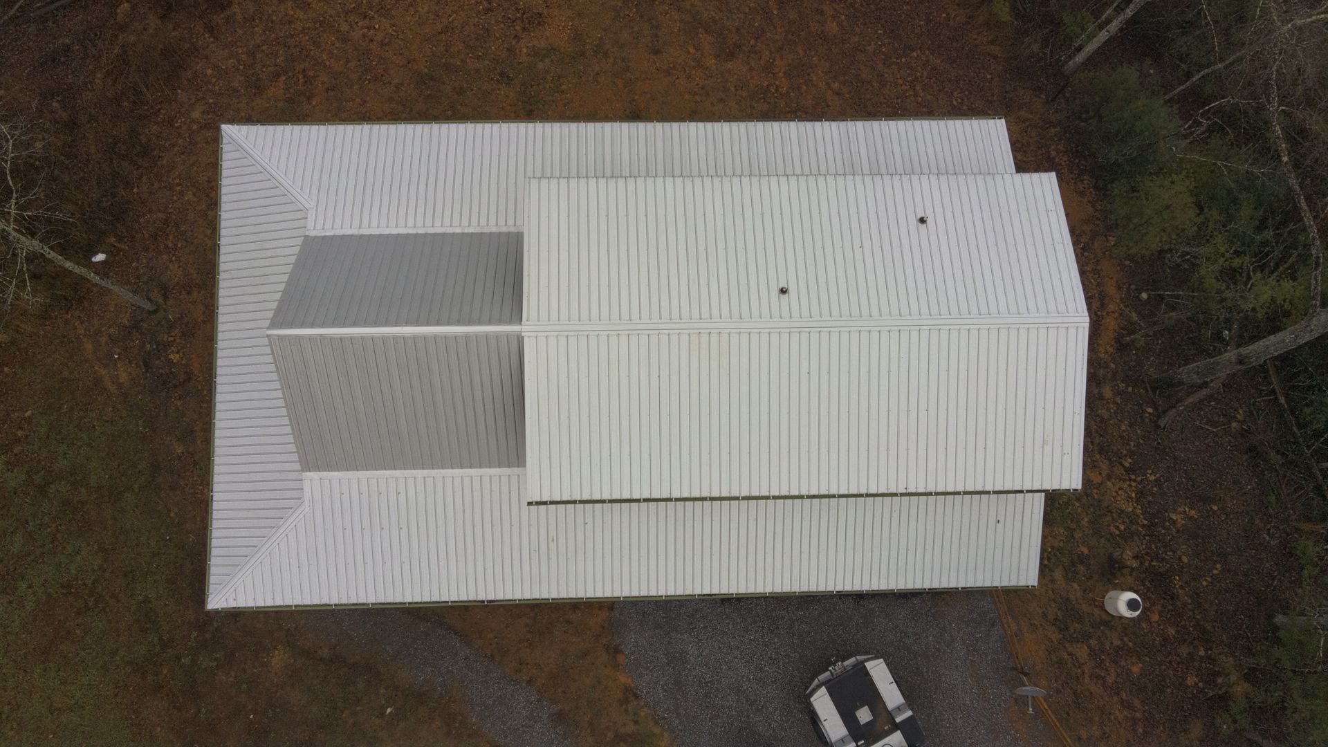 Overhead view of a white metal-roofed building in a wooded area. A vehicle is parked in the driveway.