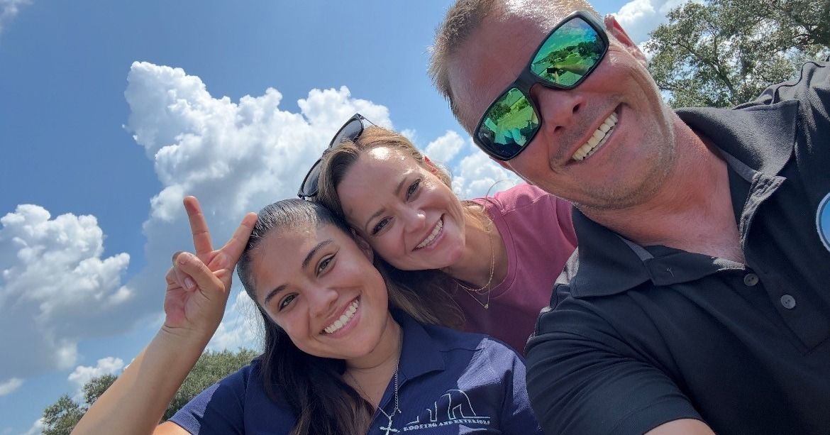 Three smiling people taking a selfie outdoors under a blue sky with fluffy clouds.