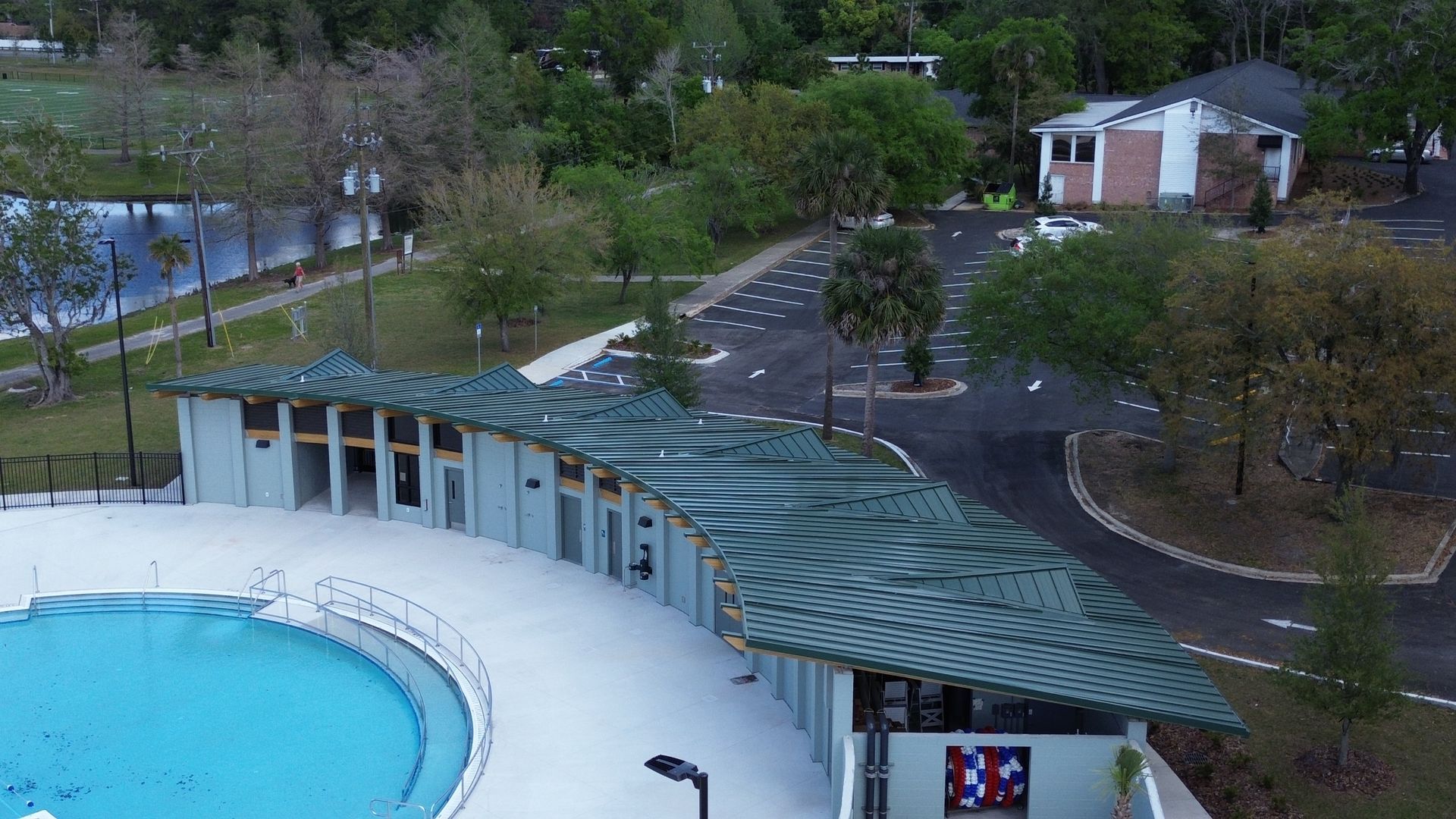 Aerial view of a swimming pool with a curved building featuring a green roof, next to a parking lot and green spaces.