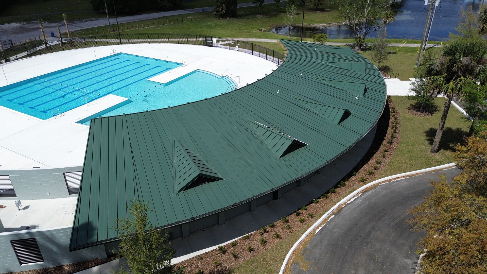 Green-roofed building curves alongside a blue rectangular swimming pool. A paved road and landscaping are also visible.