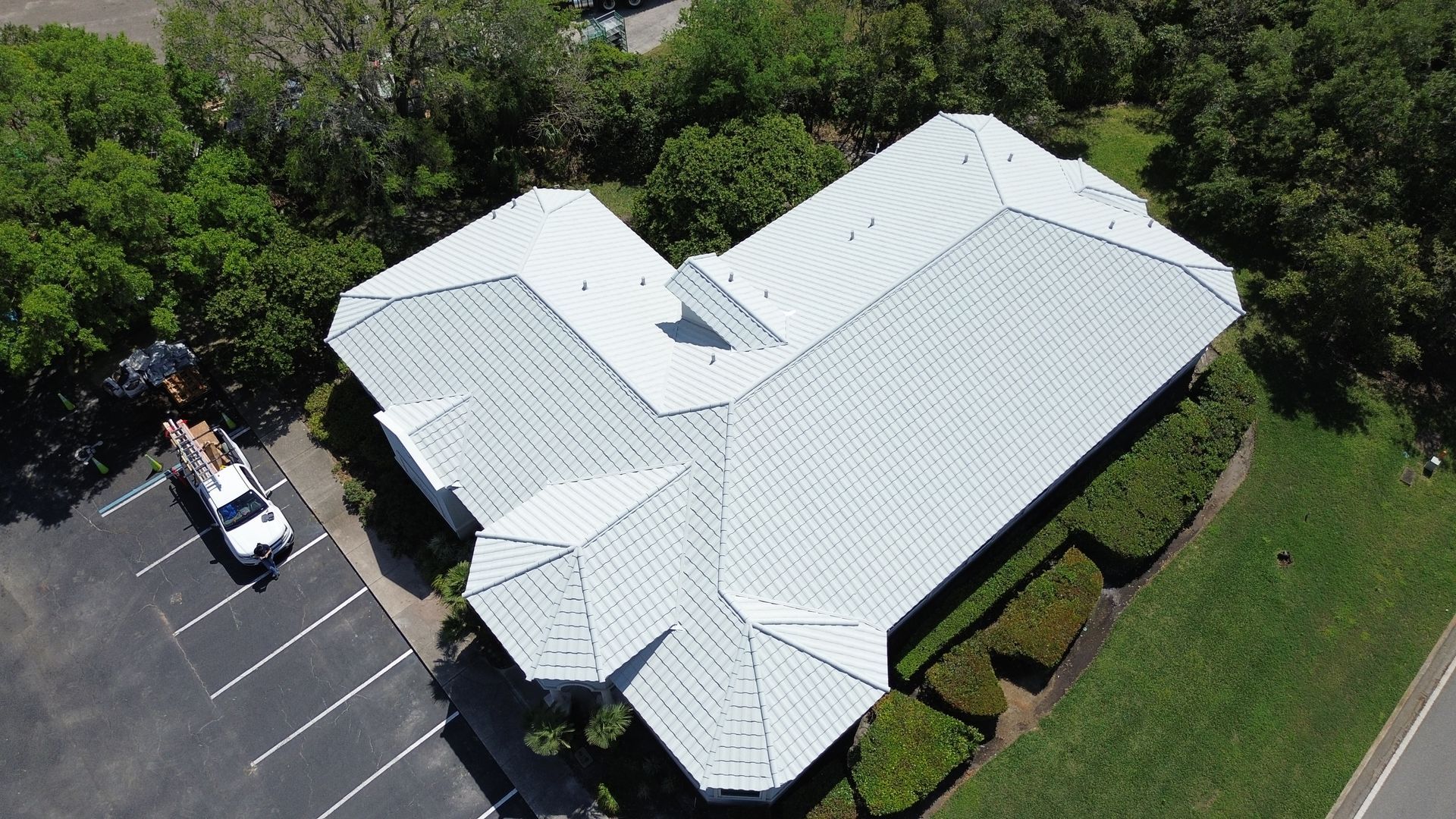 Aerial view of a building with a white metal roof, parked truck, trees, and a parking lot.