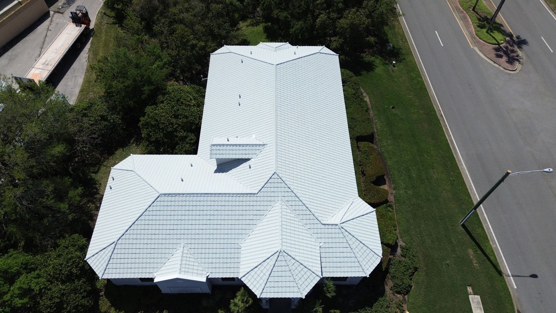Aerial view of a white-roofed house with unique angles, surrounded by trees, grass, and a road.