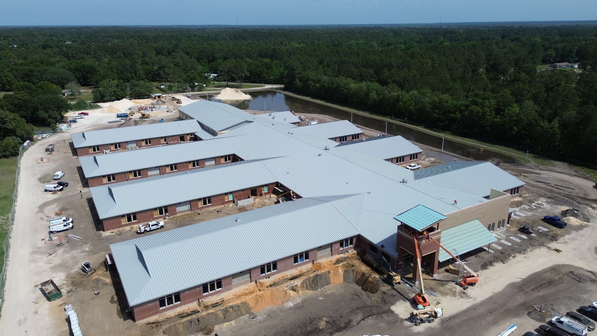 Construction site with a large, multi-winged building with a gray metal roof, surrounded by trees.