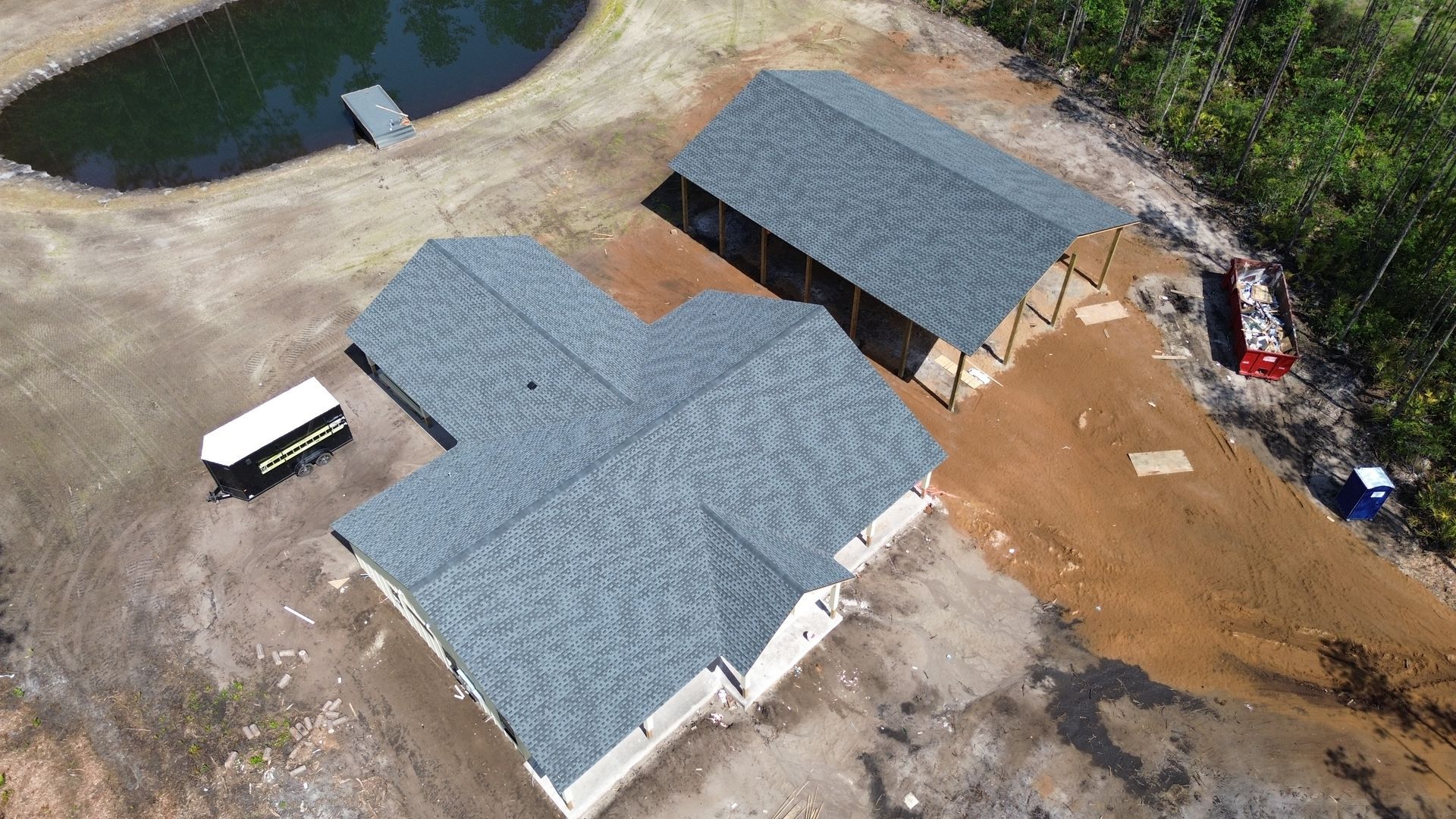 Aerial view of a house and shed with gray roofs under construction near a pond and forest.