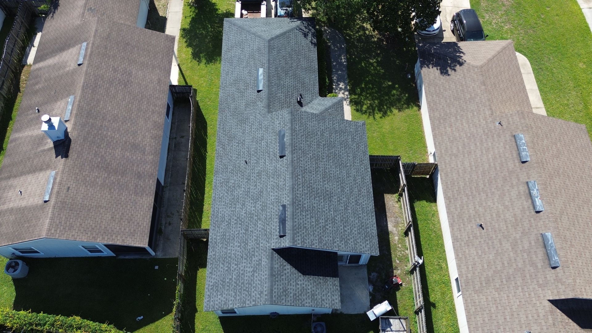 Overhead view of a house with a dark gray roof, between two other houses with brown roofs, all surrounded by green grass.