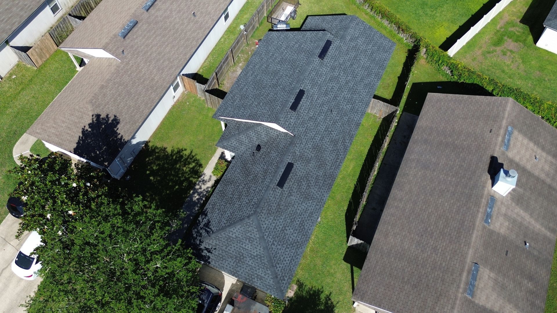 Aerial view of a house with a dark gray roof surrounded by green lawns and other houses.