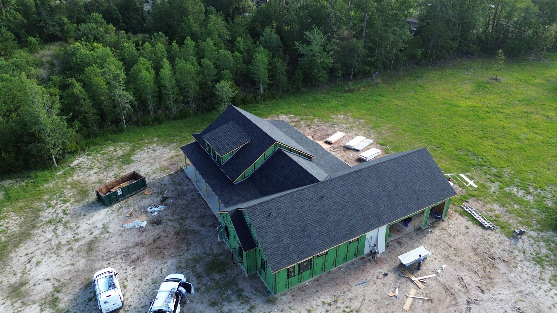 Aerial view of a house under construction with a dark roof and green framing, on a cleared lot next to trees.