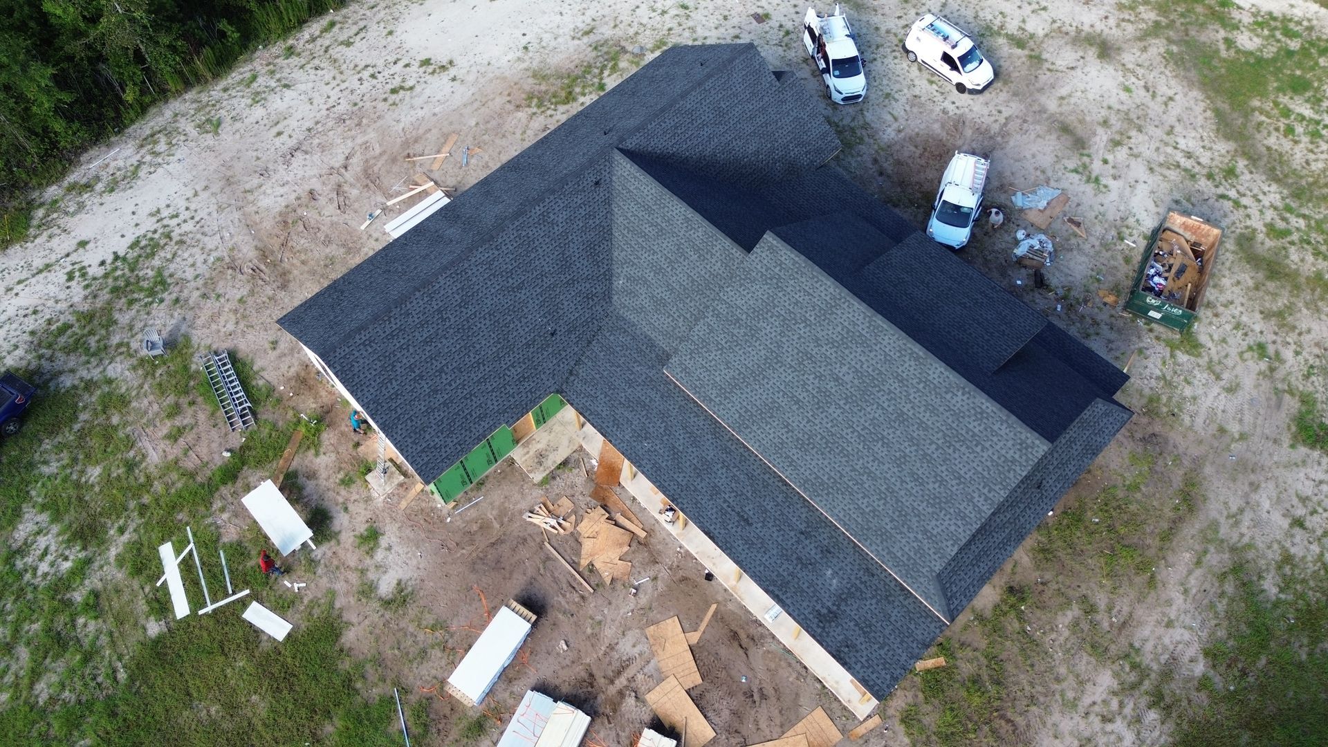 Aerial view of a partially built house with dark gray shingle roofing and construction materials on the ground.