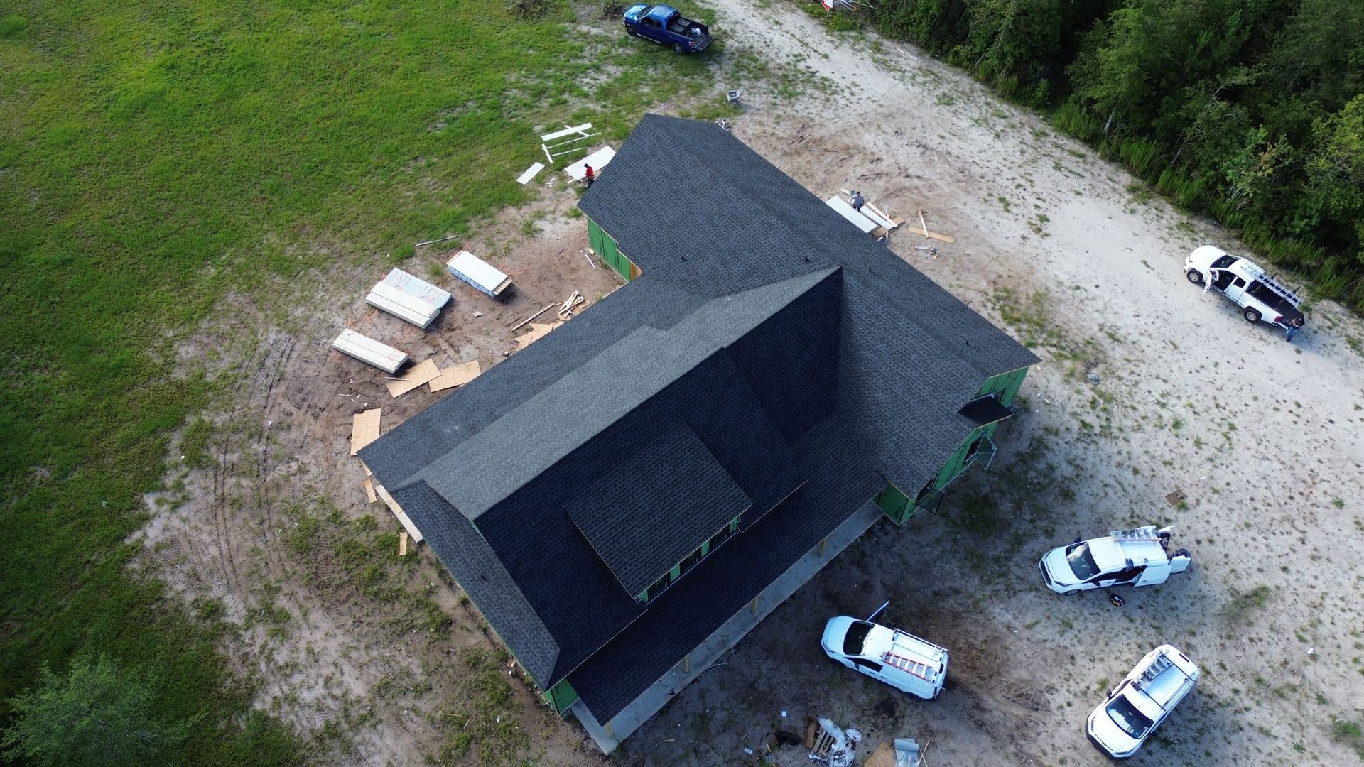 Aerial view of a dark-roofed house under construction with surrounding vehicles and materials on a sandy lot.