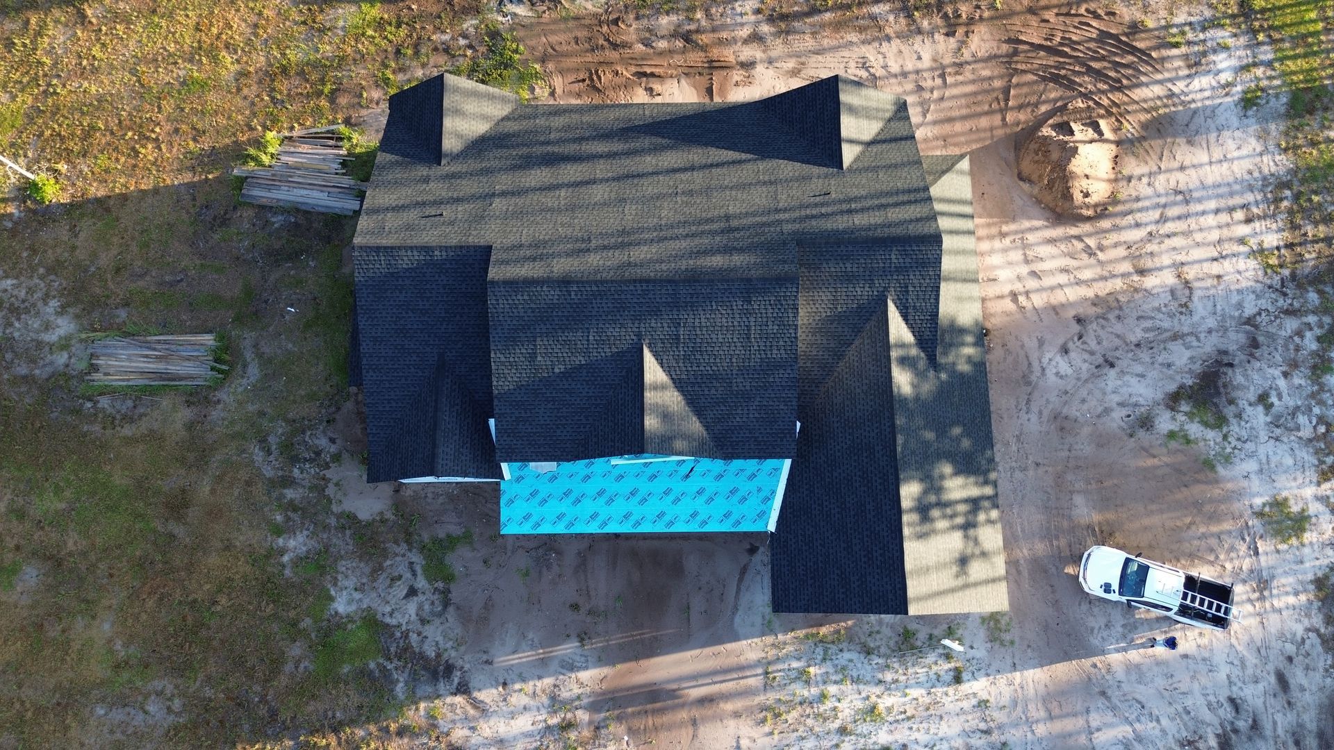 Overhead view of a house with a dark roof and a turquoise blue section, next to a white truck on a sandy area.