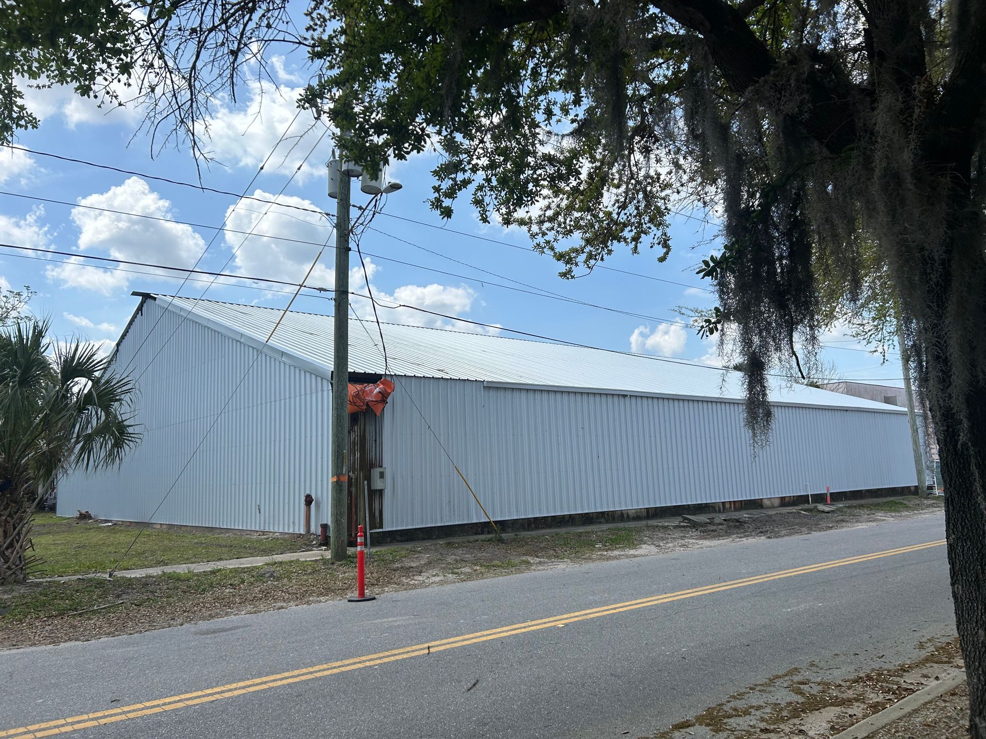 White metal warehouse building beside a road with power lines and trees overhead.